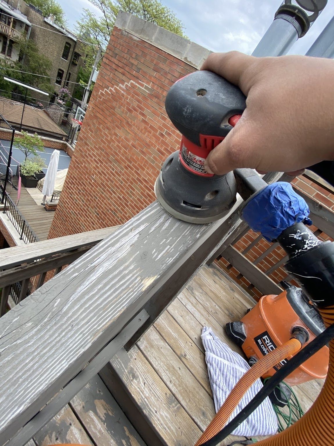 A hand uses an orbital sander to strip weathered gray paint from a wooden deck railing.
