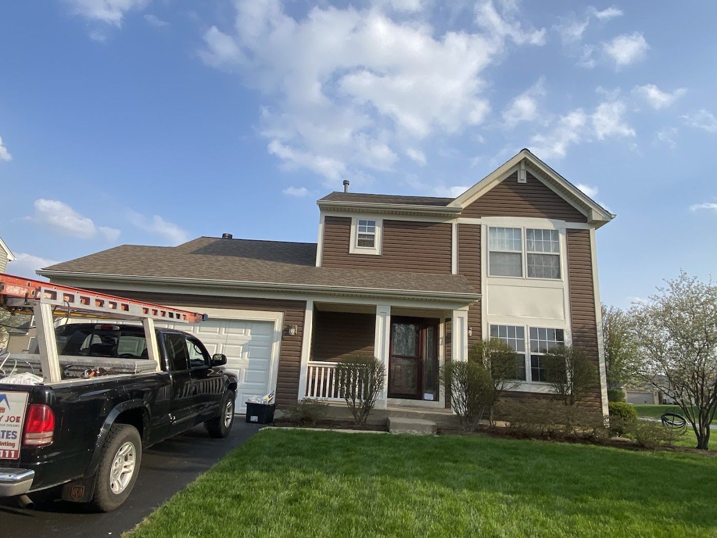 A two-story, brown wood-sided house with a black pickup truck parked in the driveway on a sunny day.