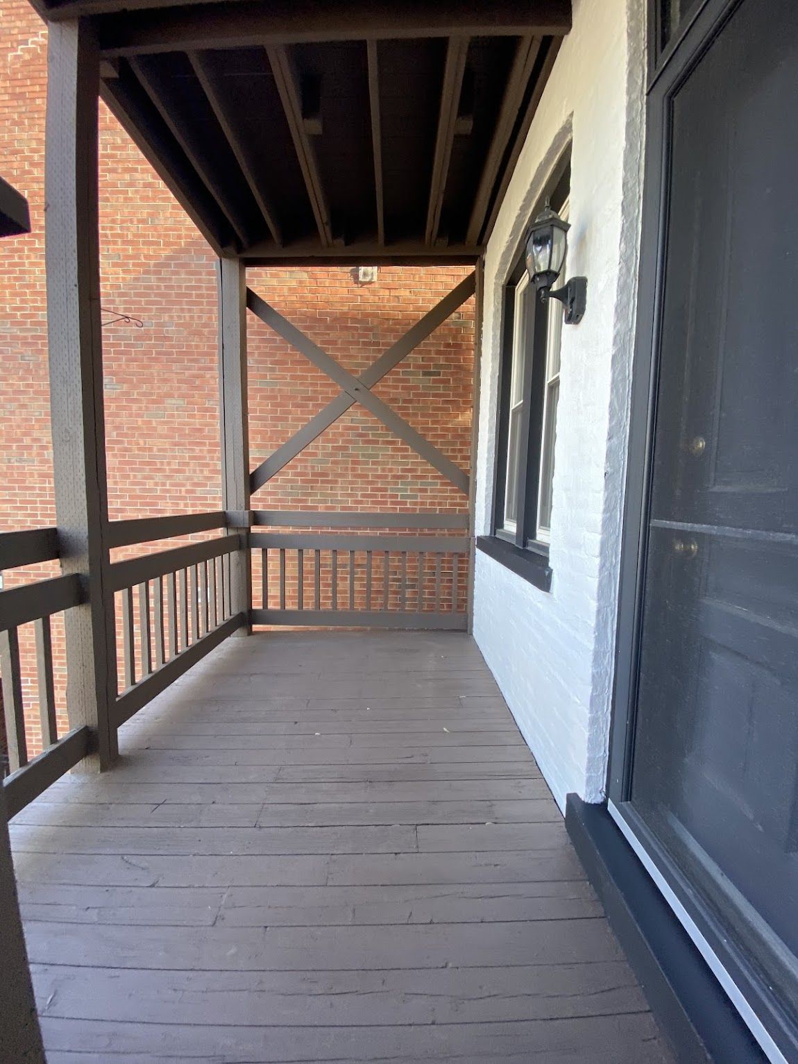A wooden porch with a railing and X-braced frame, attached to a building with white brick walls and a dark door.