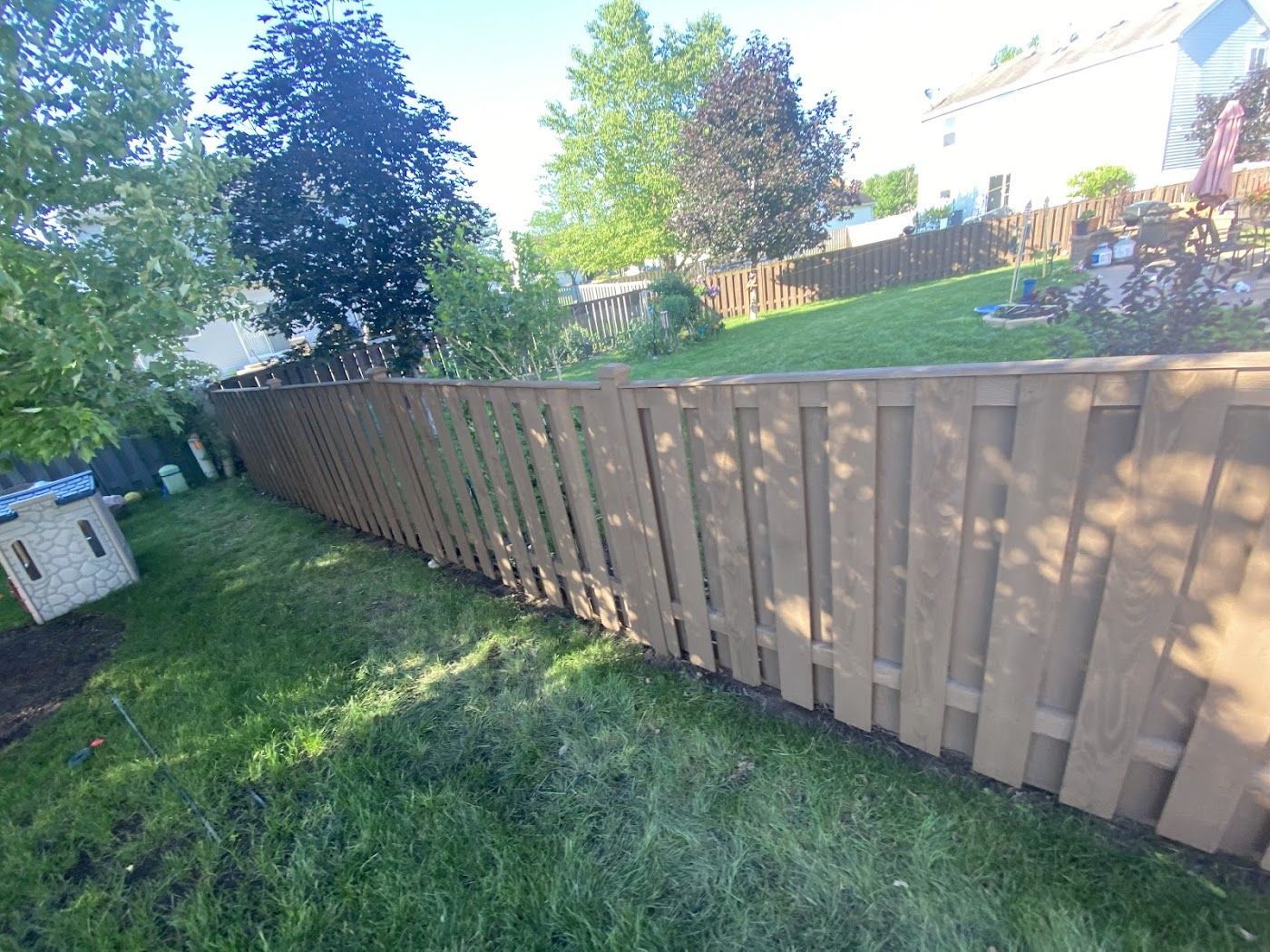 A long, wooden privacy fence runs across a grassy backyard under a bright sky with trees in the background.