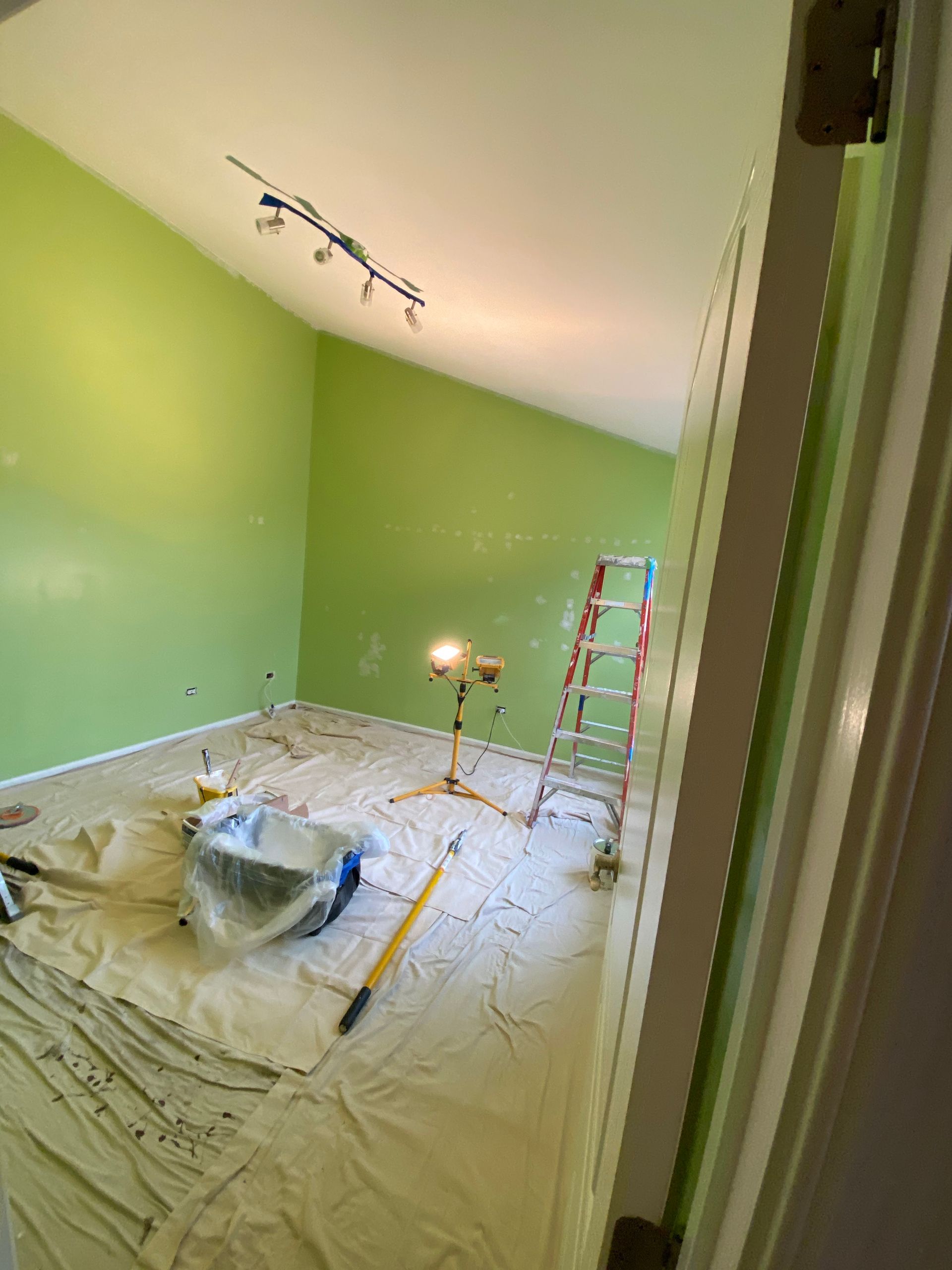 A room with light green walls under renovation, featuring a ladder, work light, and drop cloth on the floor.