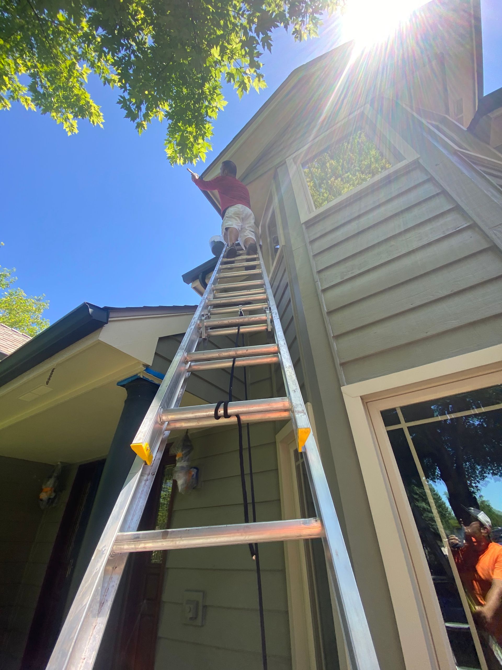 A person wearing a red shirt and light pants stands on a tall extension ladder reaching toward the roof of a house.