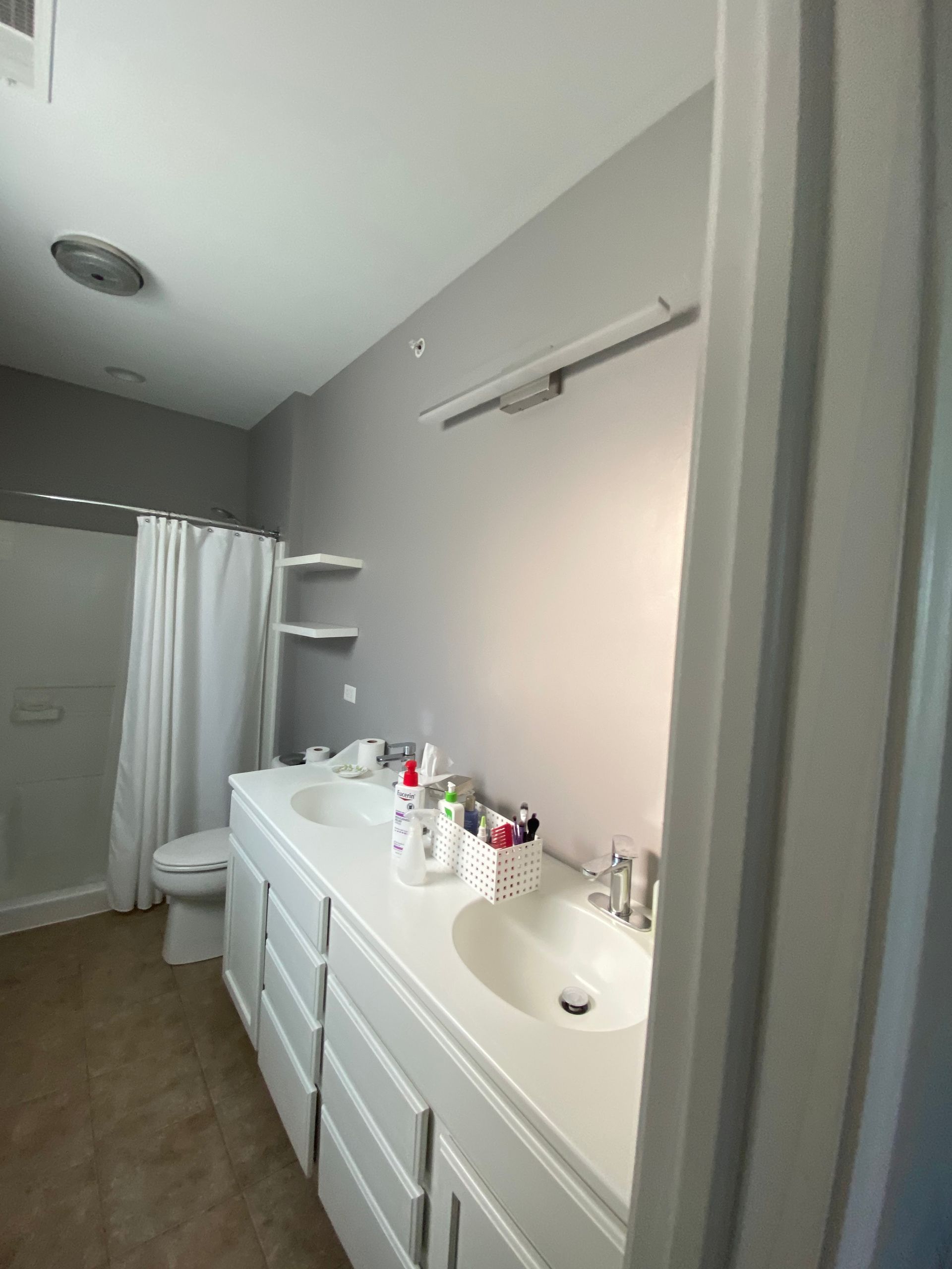 A bathroom with a white double-sink vanity, gray walls, a shower with a white curtain, and beige tile flooring.