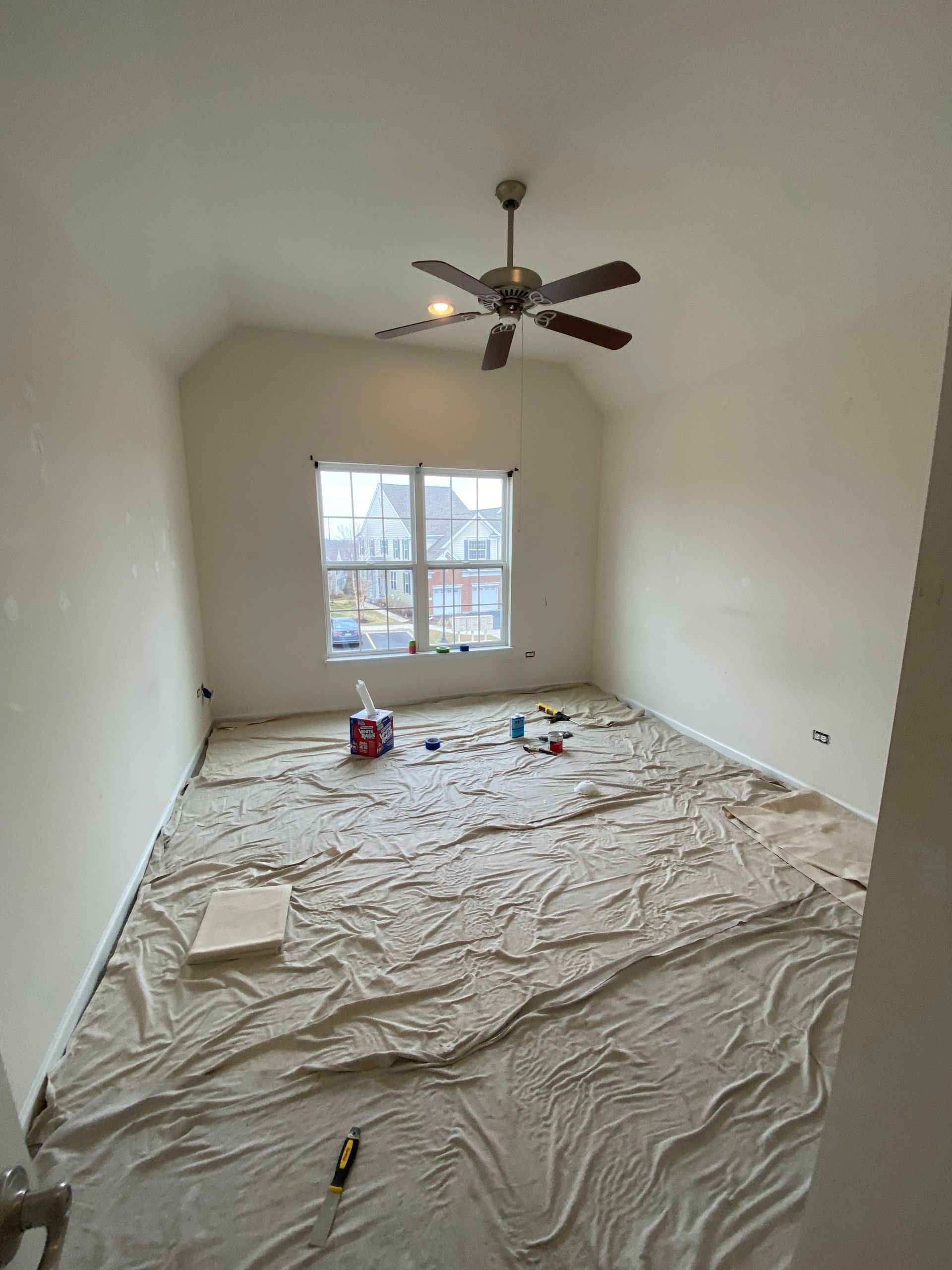 A room with light-colored walls and a ceiling fan, with a large drop cloth covering the entire floor for renovation.