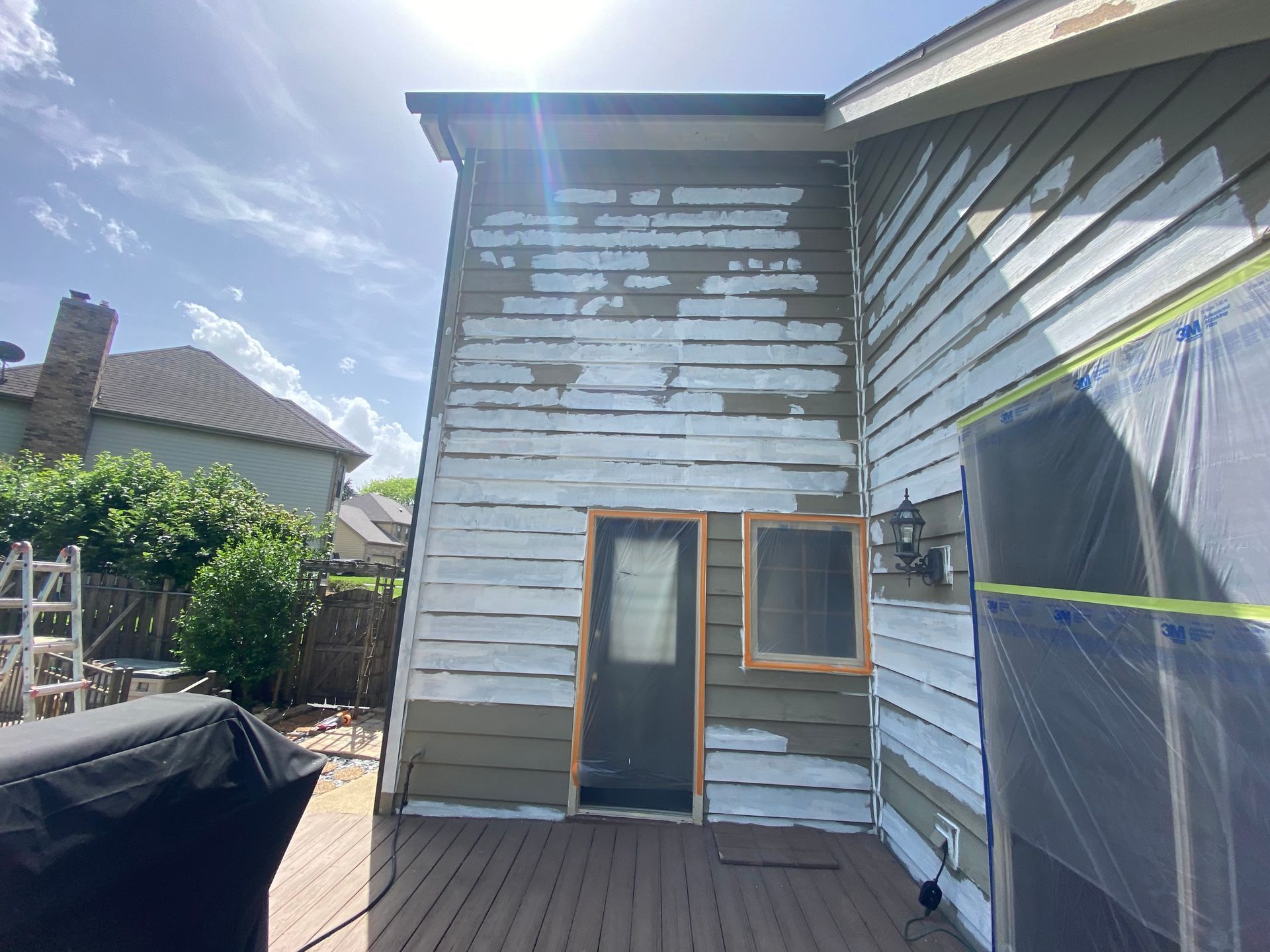 Exterior wall of a house with partially applied white primer over brown siding, next to a deck and a covered structure.