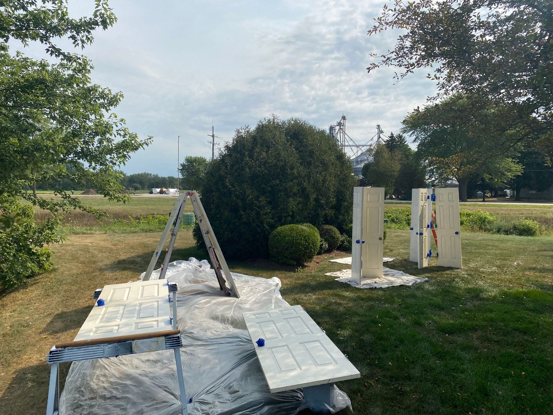 Several light-colored doors sit on a drop cloth and stand upright outdoors for painting on a sunny day.
