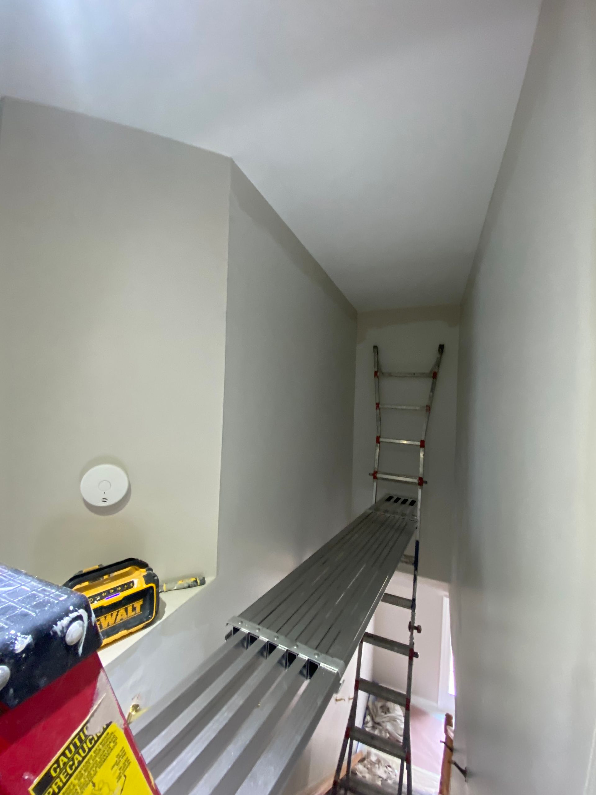 A metal scaffolding plank bridged between two ladders in a narrow hallway to reach high ceiling areas for painting.