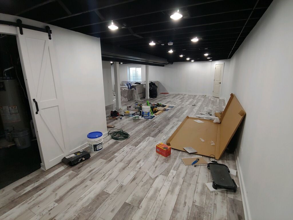 A basement room under renovation with white walls, black ceiling, wood-look flooring, a barn door, and scattered supplies.