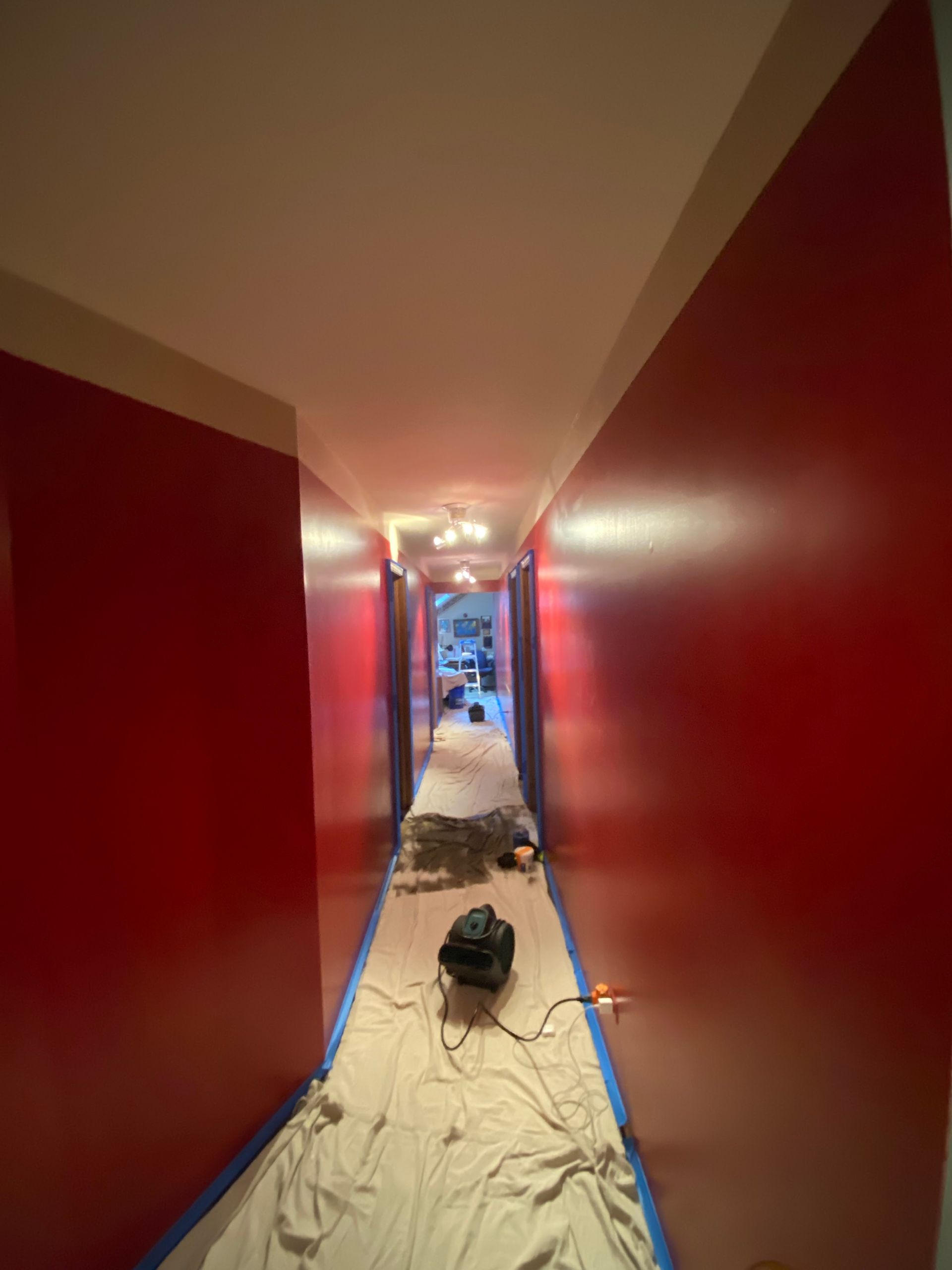 A narrow hallway with freshly painted deep red walls, floor covered in drop cloths, and a small fan in the center.