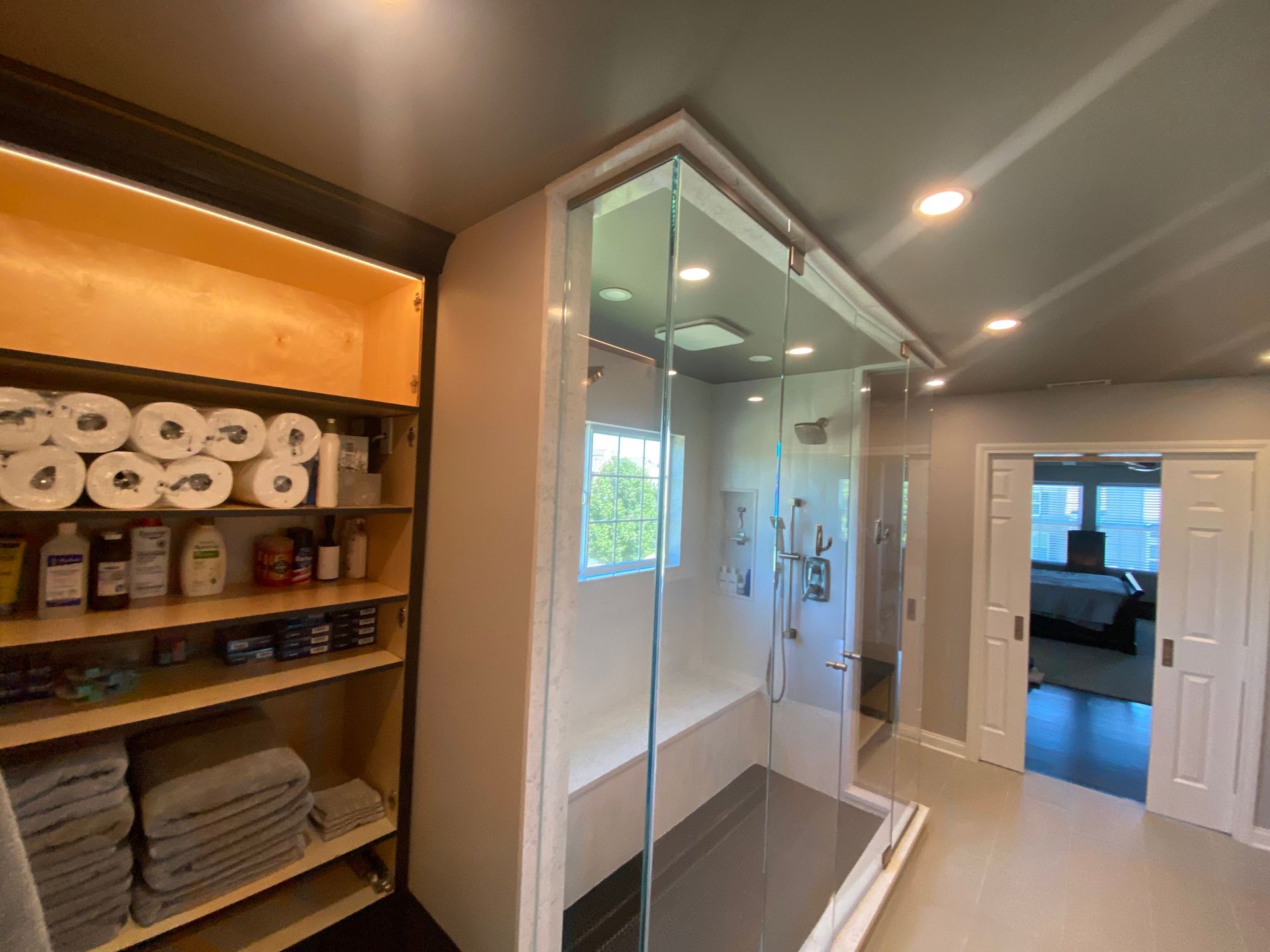 A modern bathroom featuring built-in wooden shelves stocked with towels and toiletries beside a glass walk-in shower.