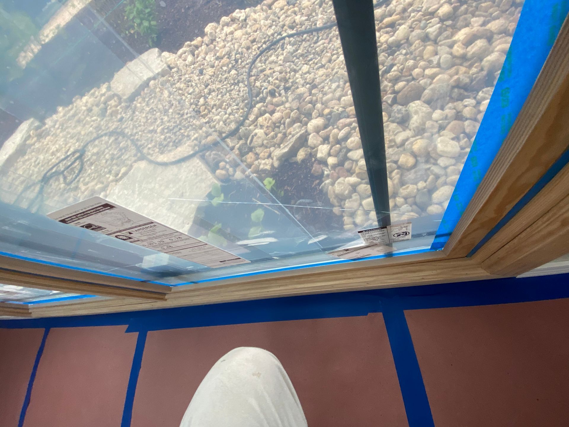 A view through a window covered in protective film, looking out onto a gravel bed, with blue painter's tape on the sill.