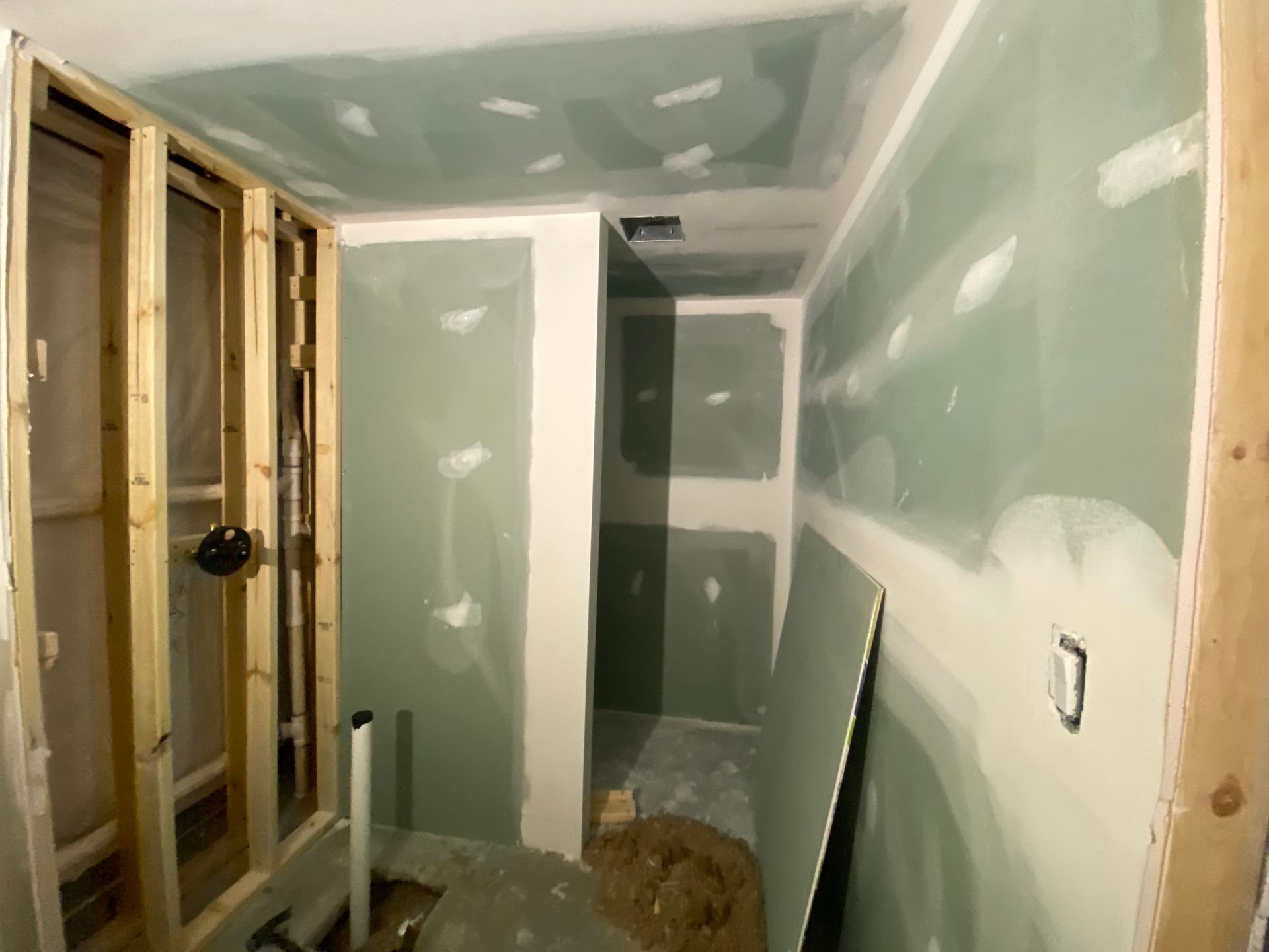 An interior view of a room under construction with green moisture-resistant drywall, wooden wall framing, and a pipe.