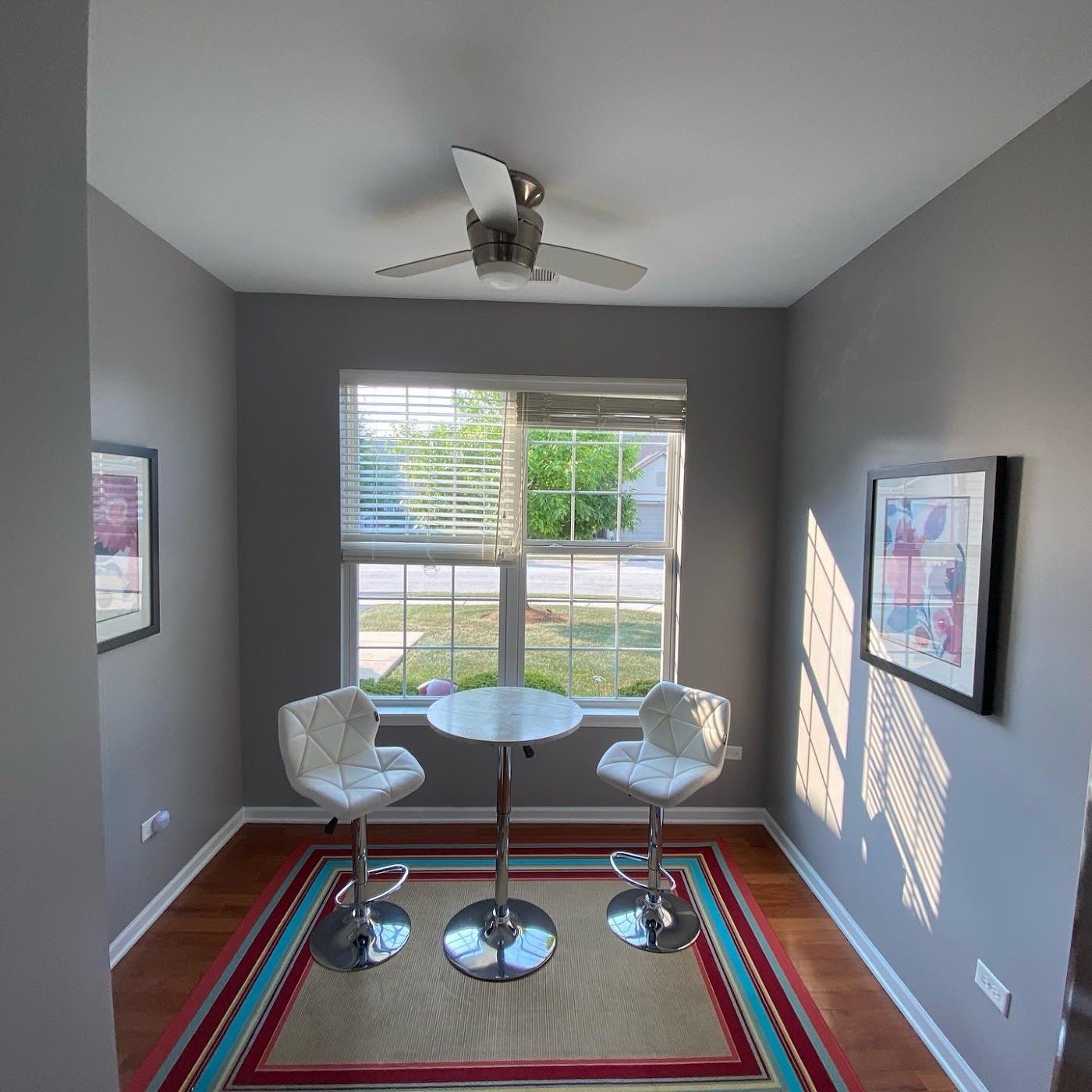 A small bistro table with two white stools sits on a colorful rug in a room with grey walls and a ceiling fan.