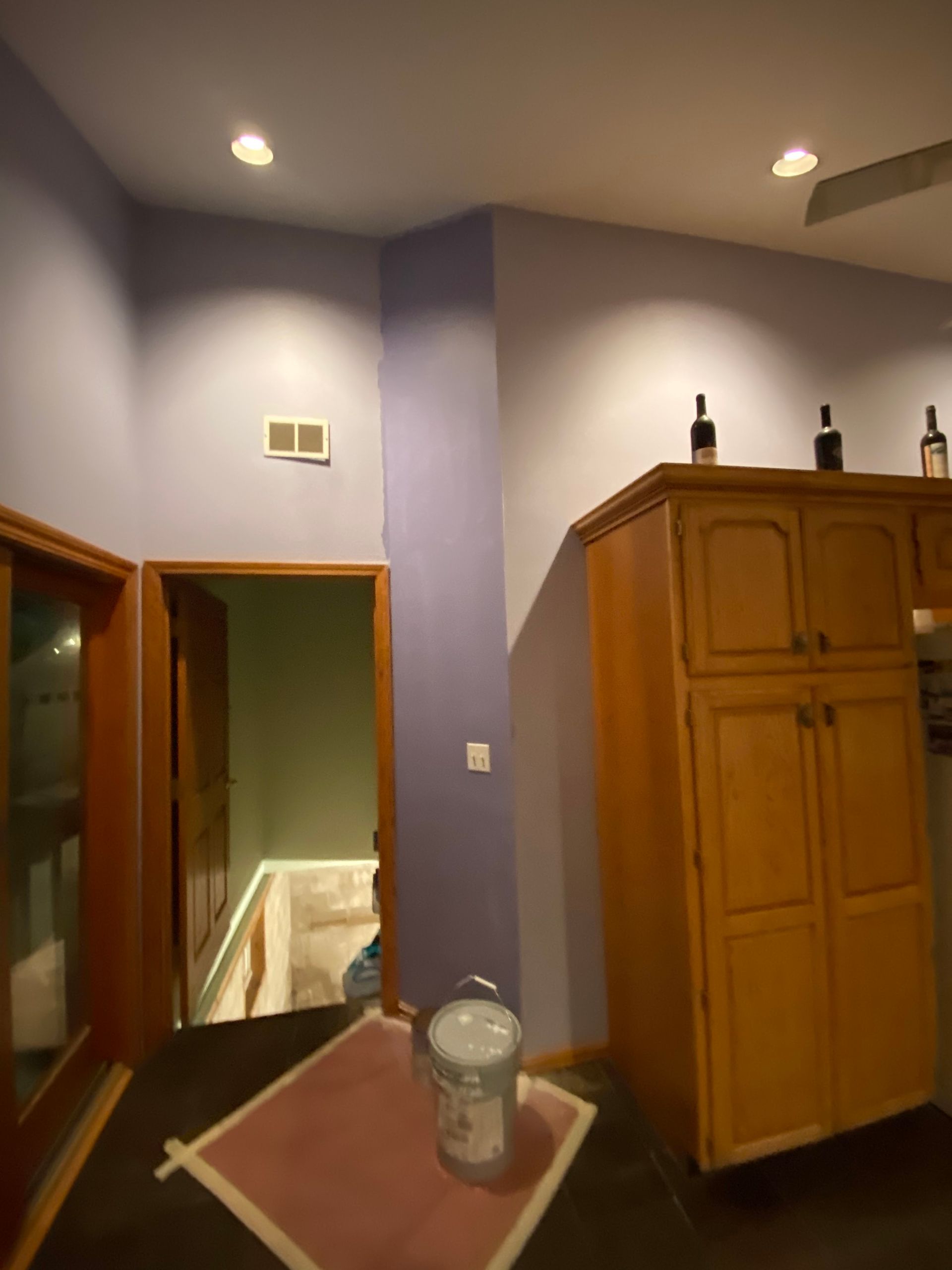 A room with light purple walls, wooden cabinets, a doorway to a stairwell, recessed lighting, and a paint bucket on a mat.