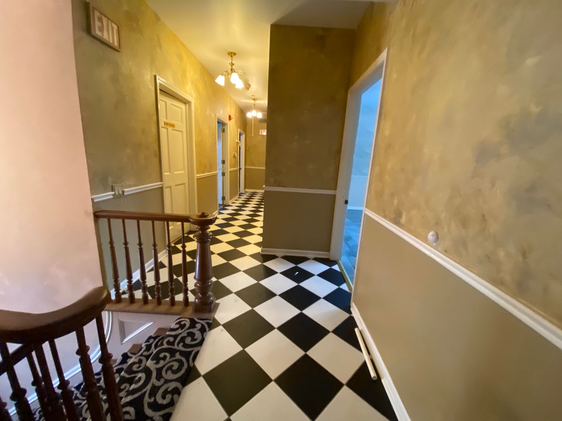 A hallway with black-and-white checkered floors, tan walls with wainscoting, and wooden railings leading to a staircase.