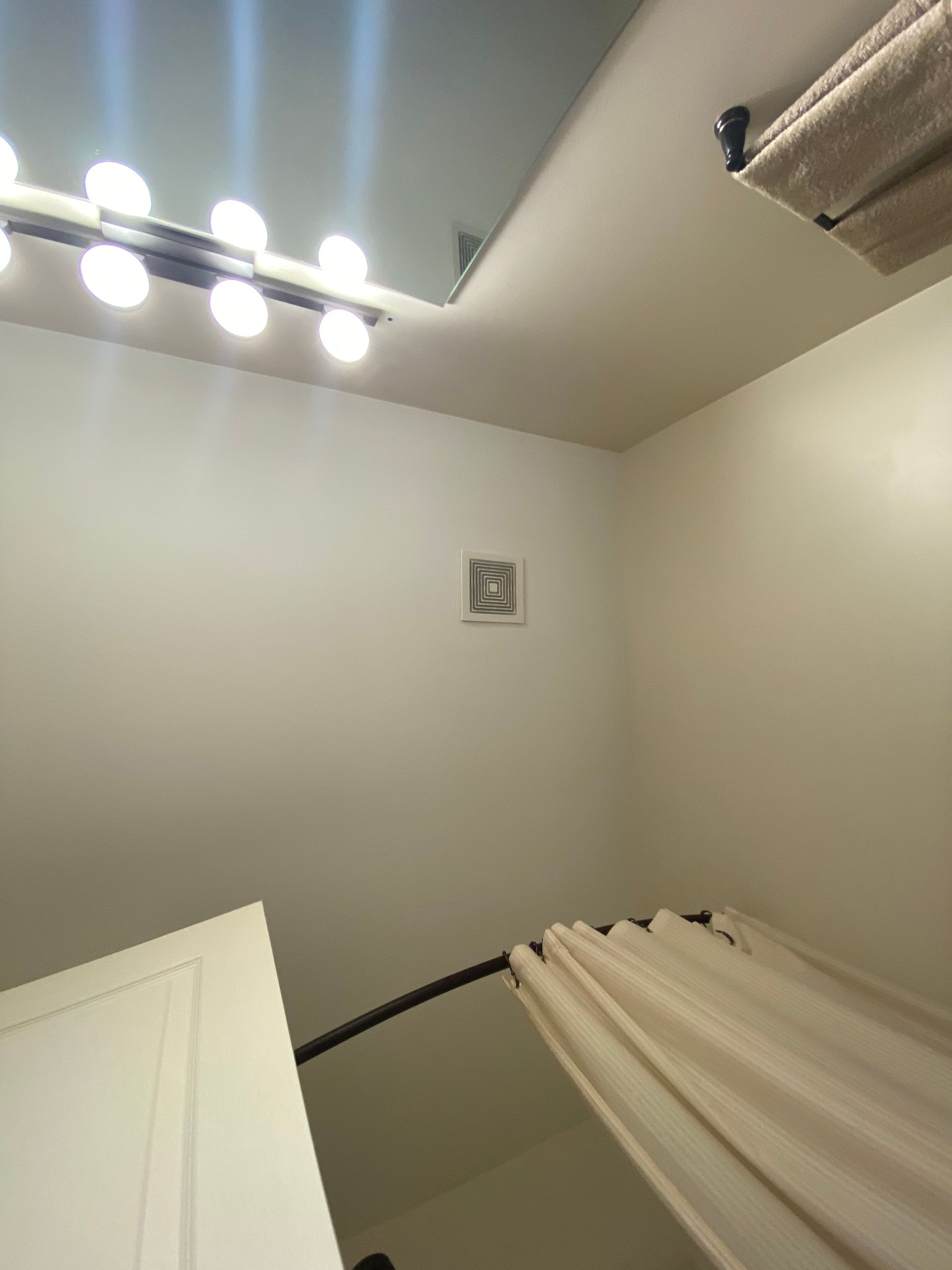 Low-angle view of a bathroom corner featuring a vanity light, a wall vent, and a shower curtain hanging on a curved rod.
