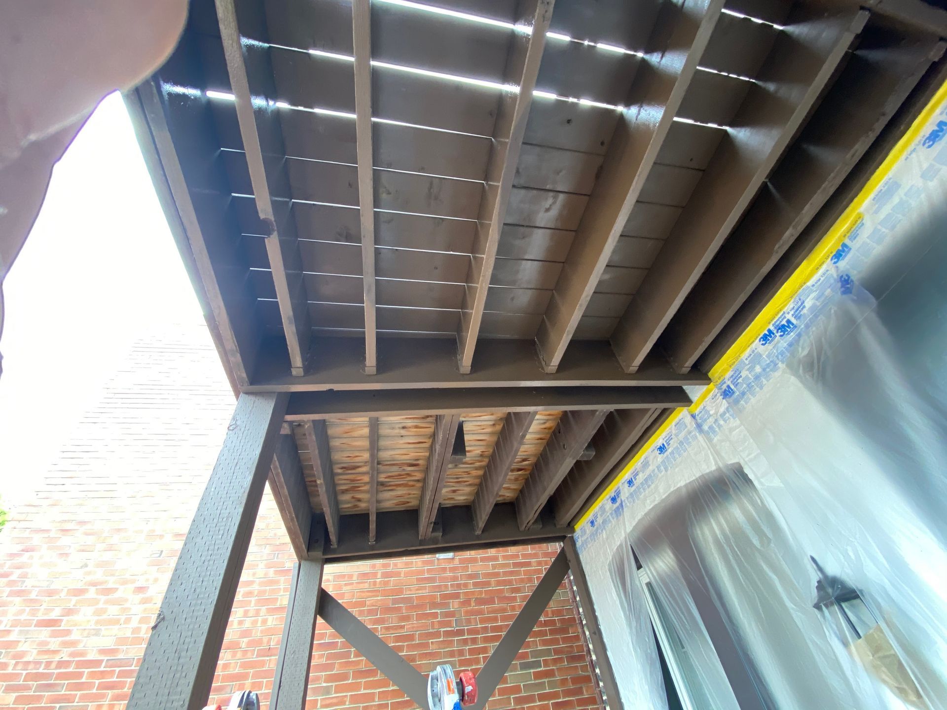 Low-angle view of a wooden deck underside showing joists, exposed plywood, and a brick wall next to a house entrance.