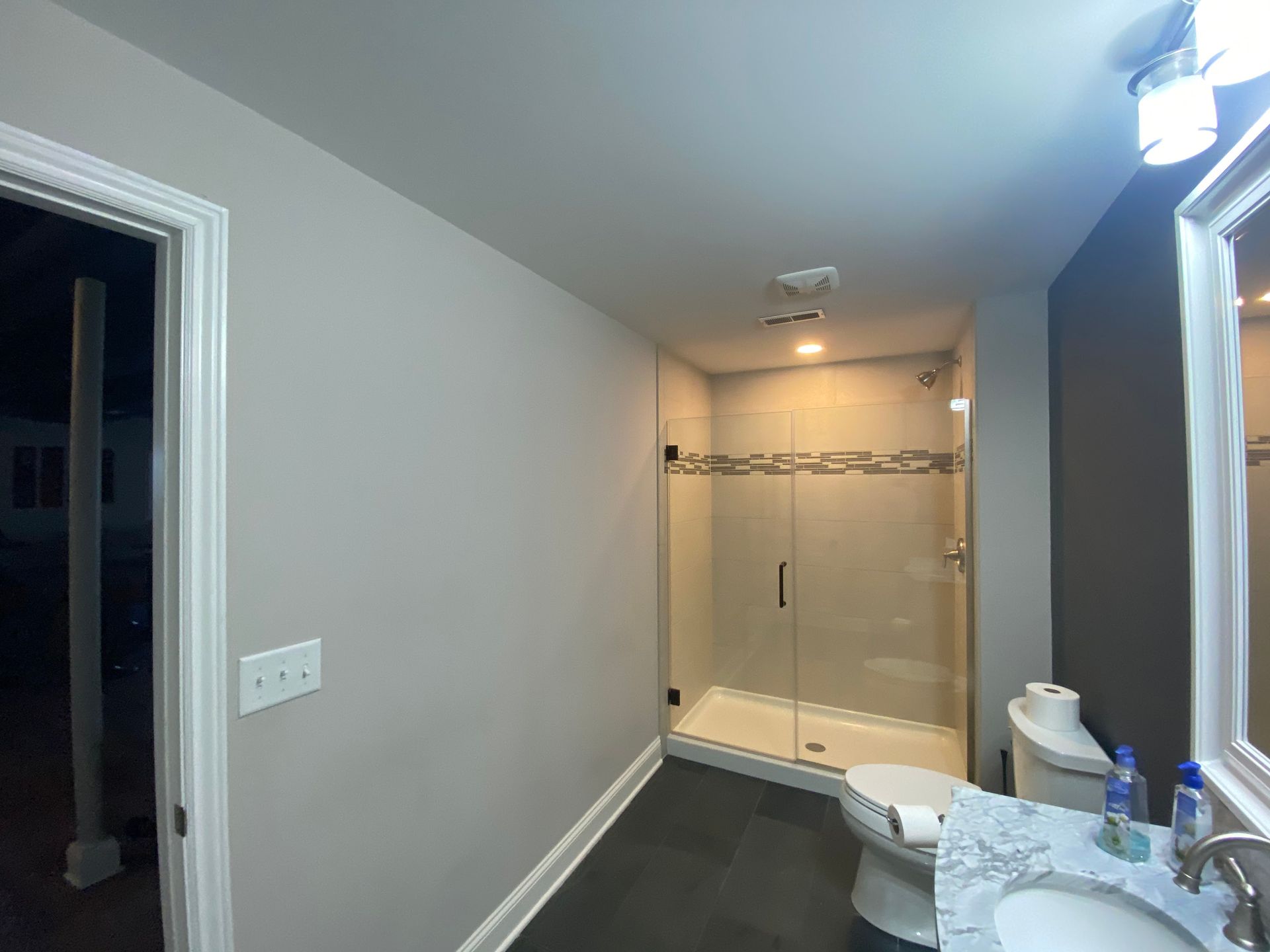 A modern bathroom with light gray walls, a glass-enclosed shower, dark flooring, and a marble-topped vanity.