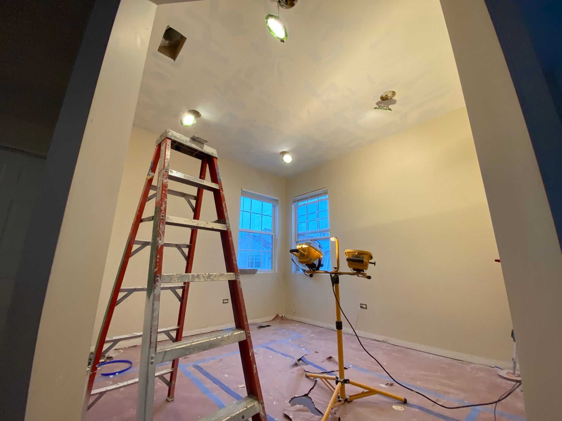 An interior room under renovation with a ladder, tripod work light, and plastic drop cloth on the floor.