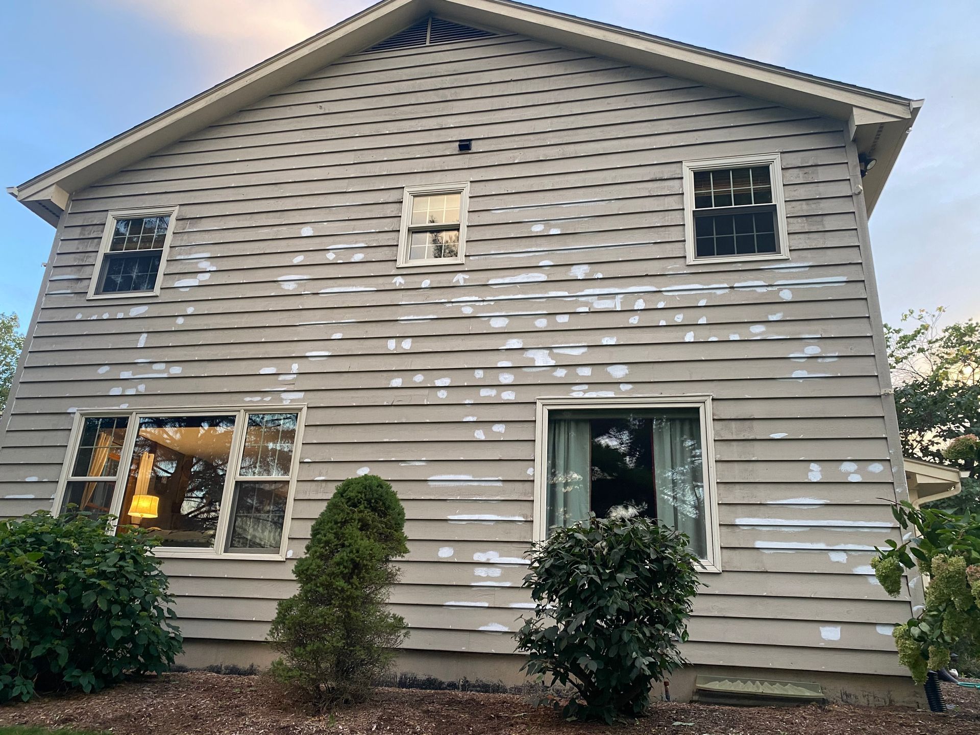 A two-story house with beige wood siding that is significantly chipped and peeling, surrounded by small green shrubs.