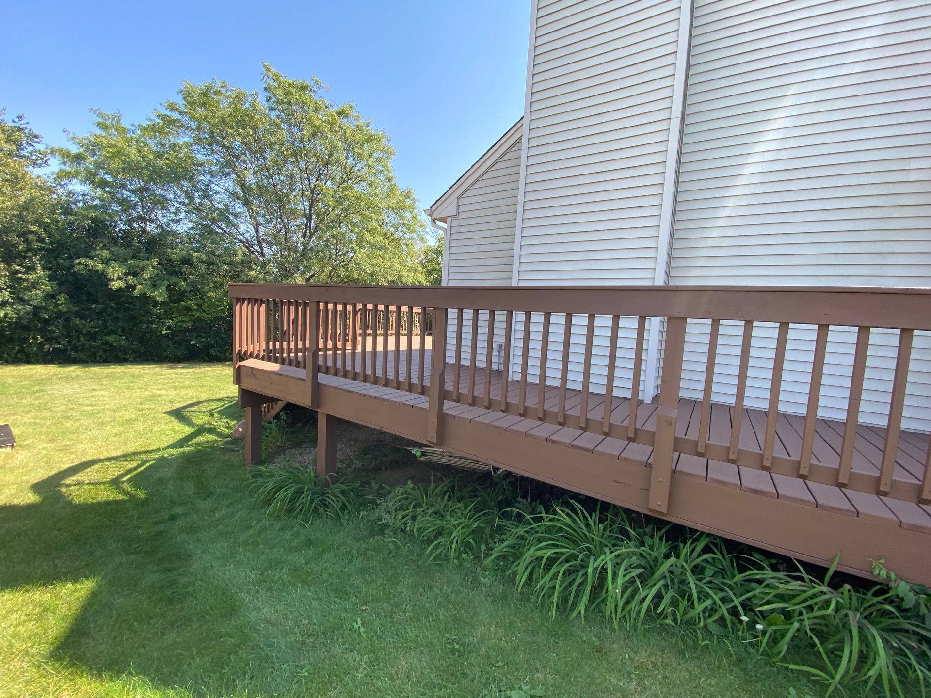A brown wooden deck attached to the side of a white house, overlooking a grassy lawn with trees in the background.