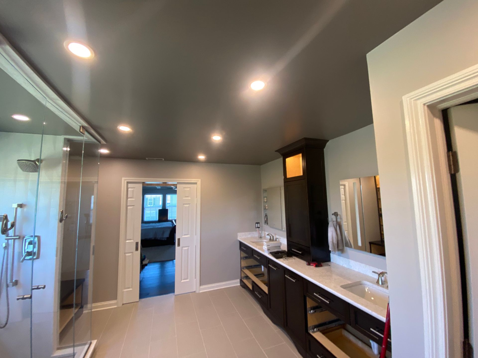 A bathroom with a dark ceiling, recessed lighting, a glass shower, dual sinks in dark cabinetry, and white sliding doors.