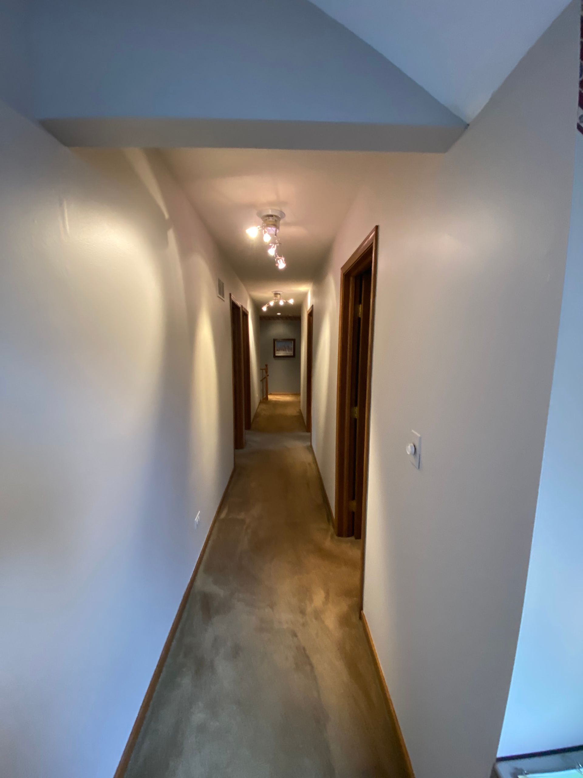 A long hallway with light-colored walls, wood-tone flooring, and multiple wooden doors, illuminated by overhead lighting.