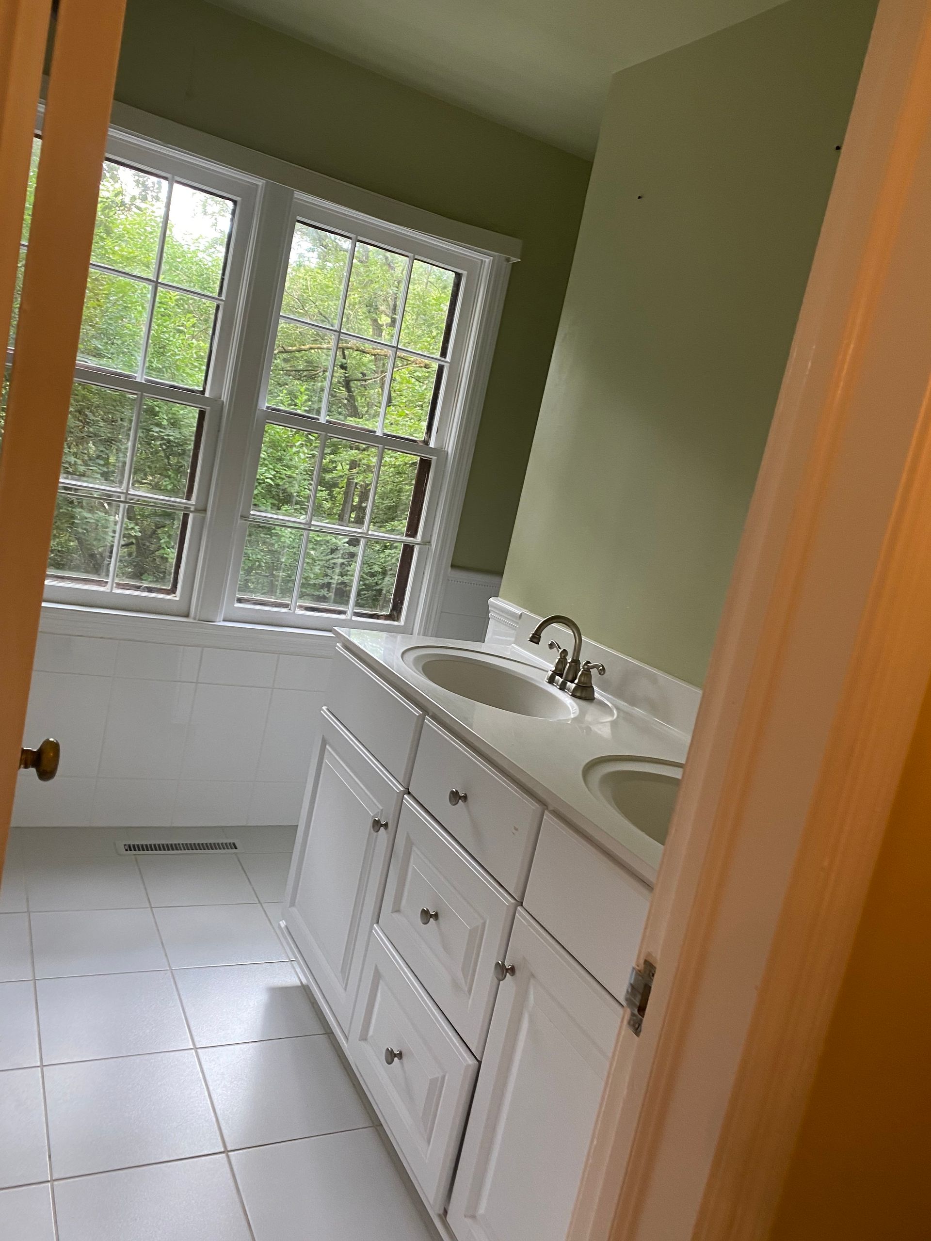 A bright bathroom featuring a white double vanity, light green walls, a window, and white tiled flooring.
