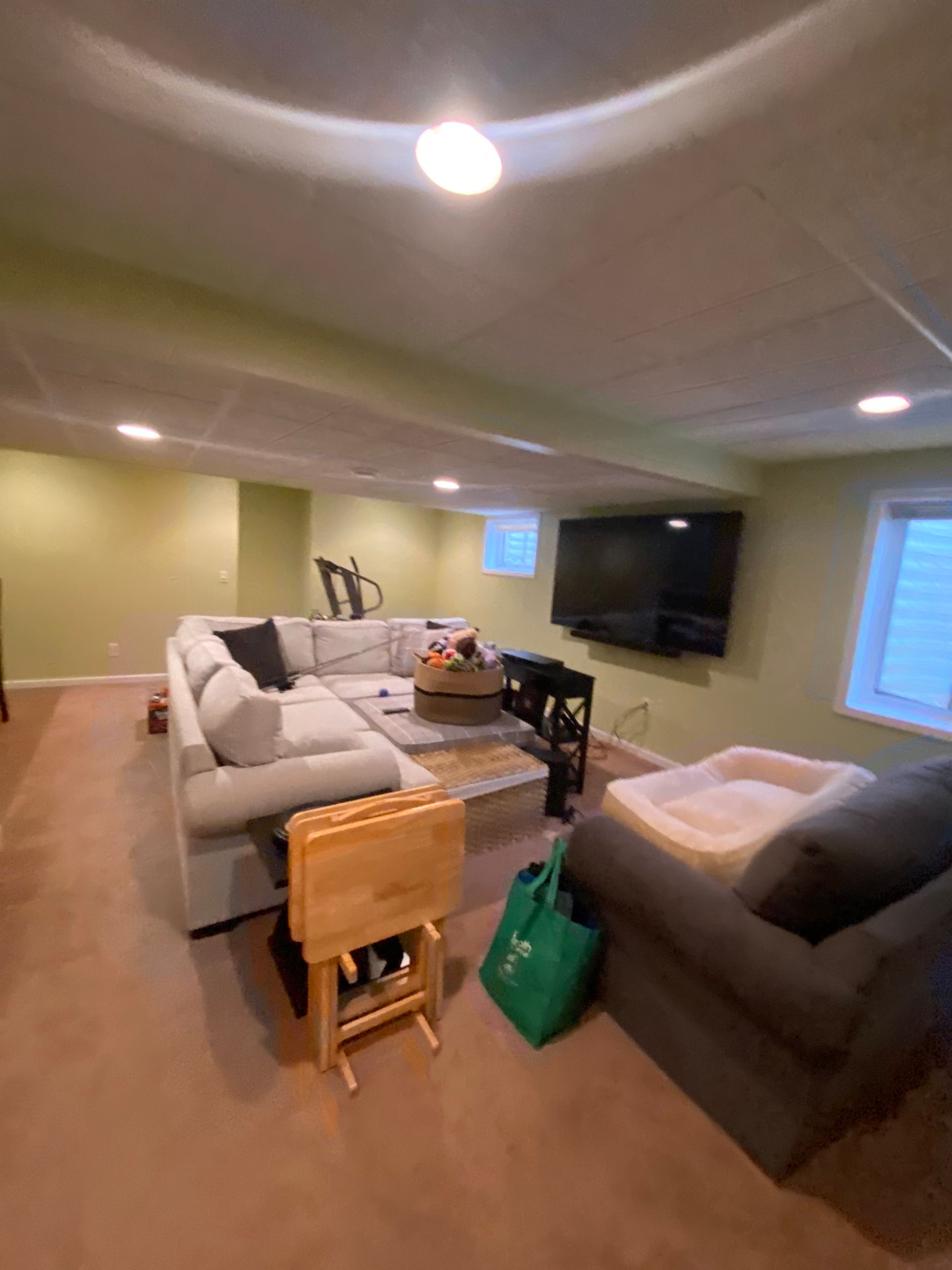 A basement living room with sage green walls, carpet, a light-colored sectional, a dark armchair, and a mounted TV.