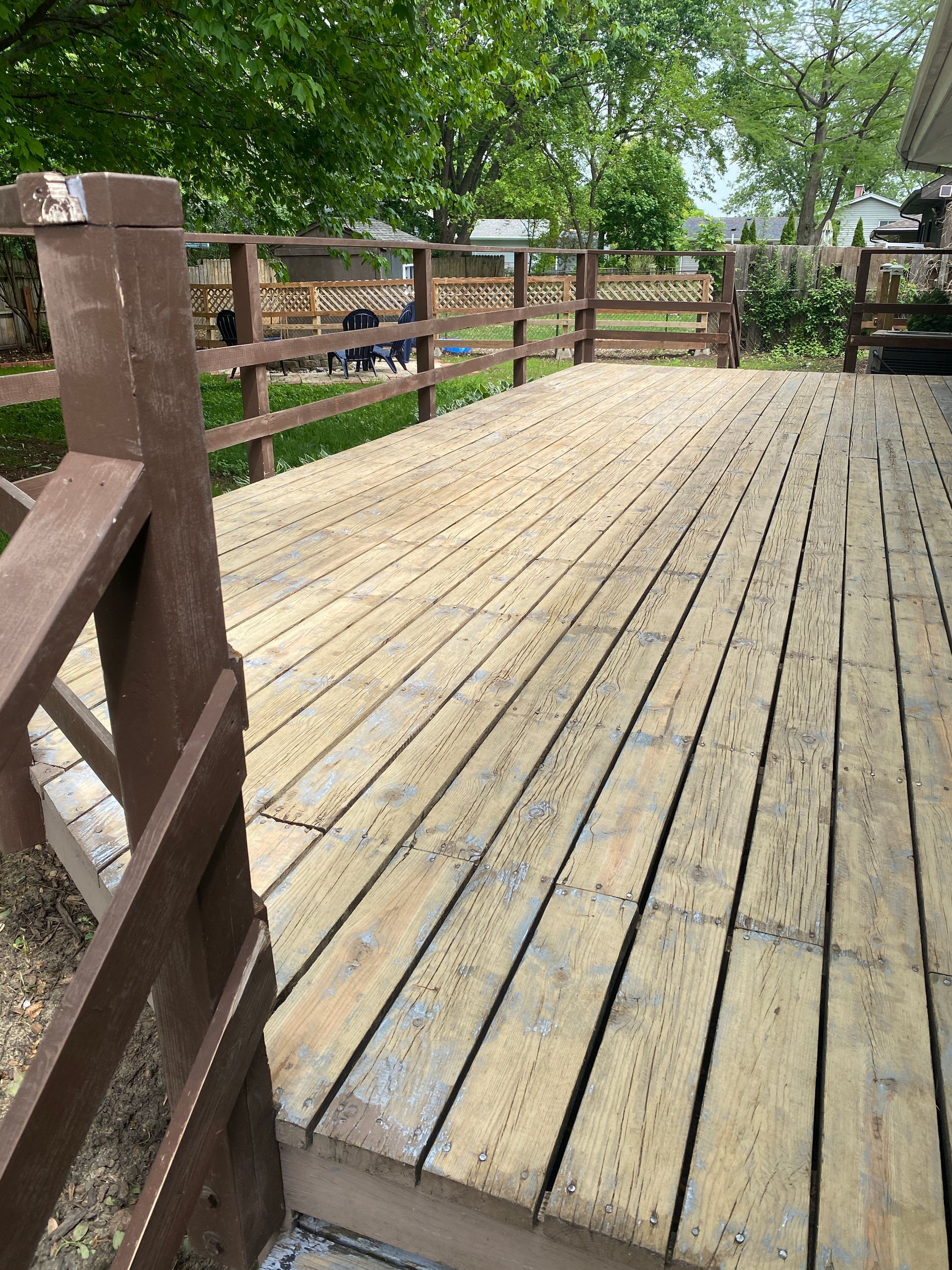 A light-colored wooden deck with brown railings, overlooking a backyard with trees and a fence on a sunny day.