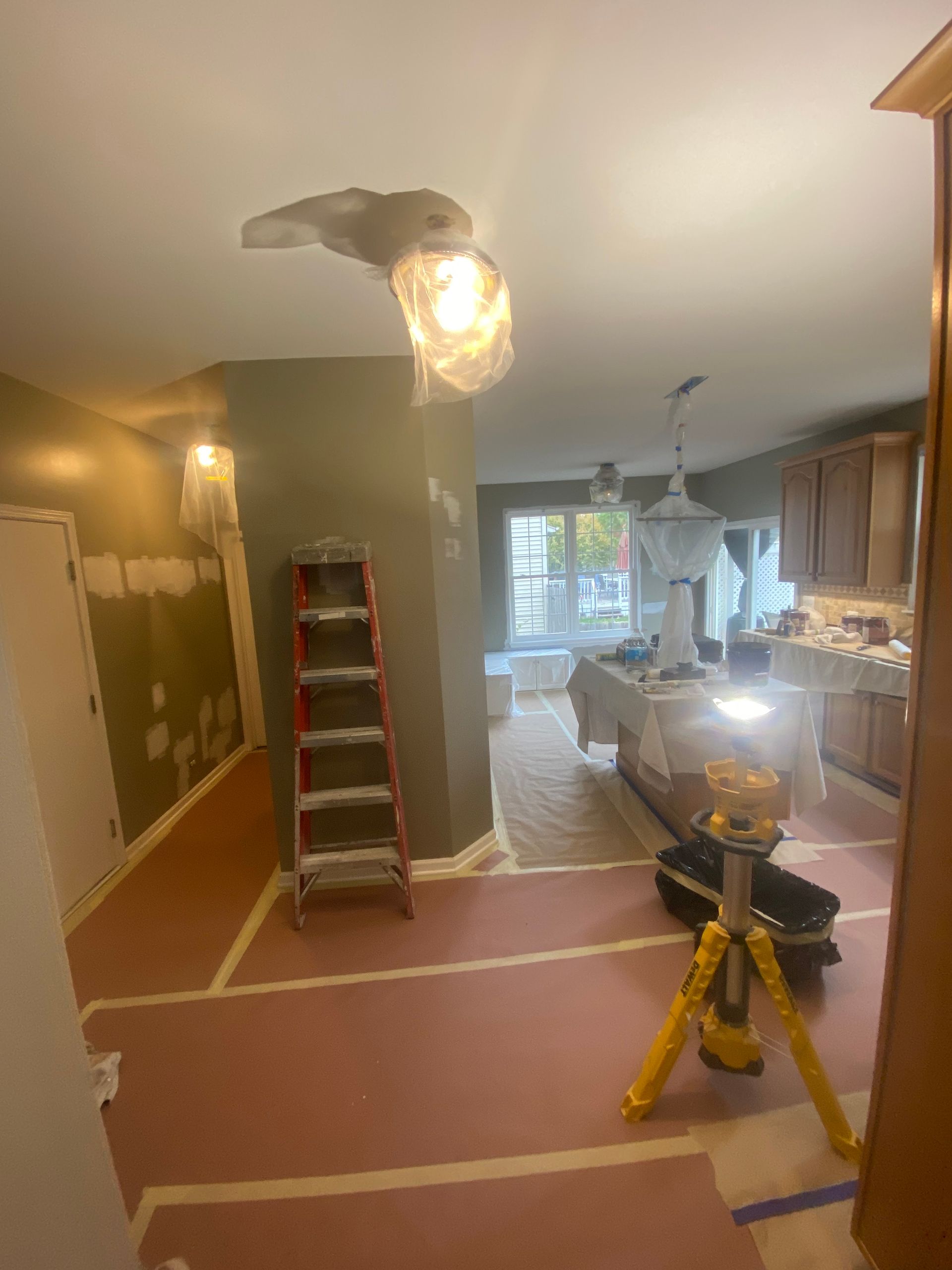 An interior view of a room under renovation, showing a ladder, work lights, floor protection paper, and taped walls.