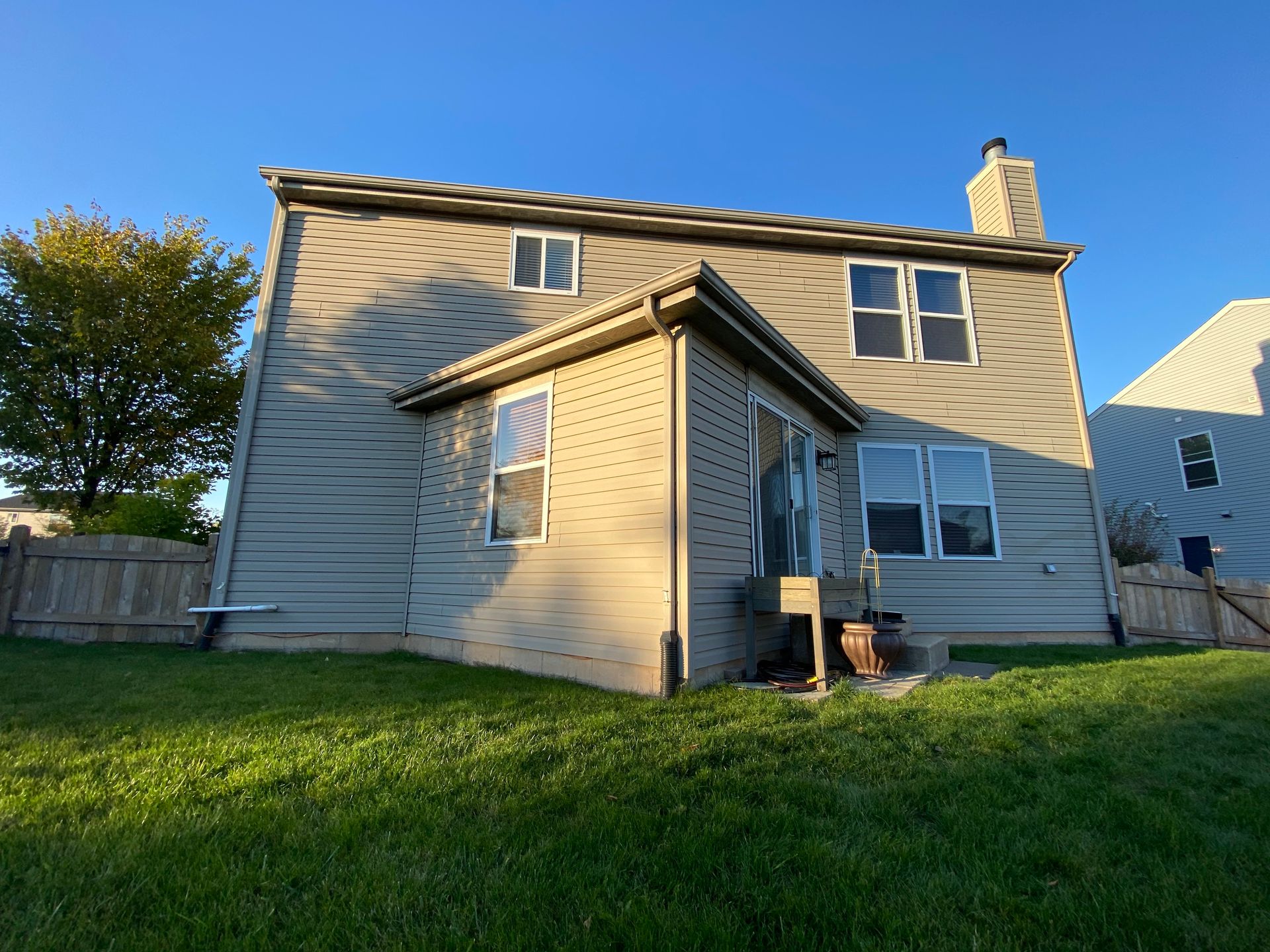 Tan two-story house with beige vinyl siding and a small attached back porch, set against a clear blue sky.