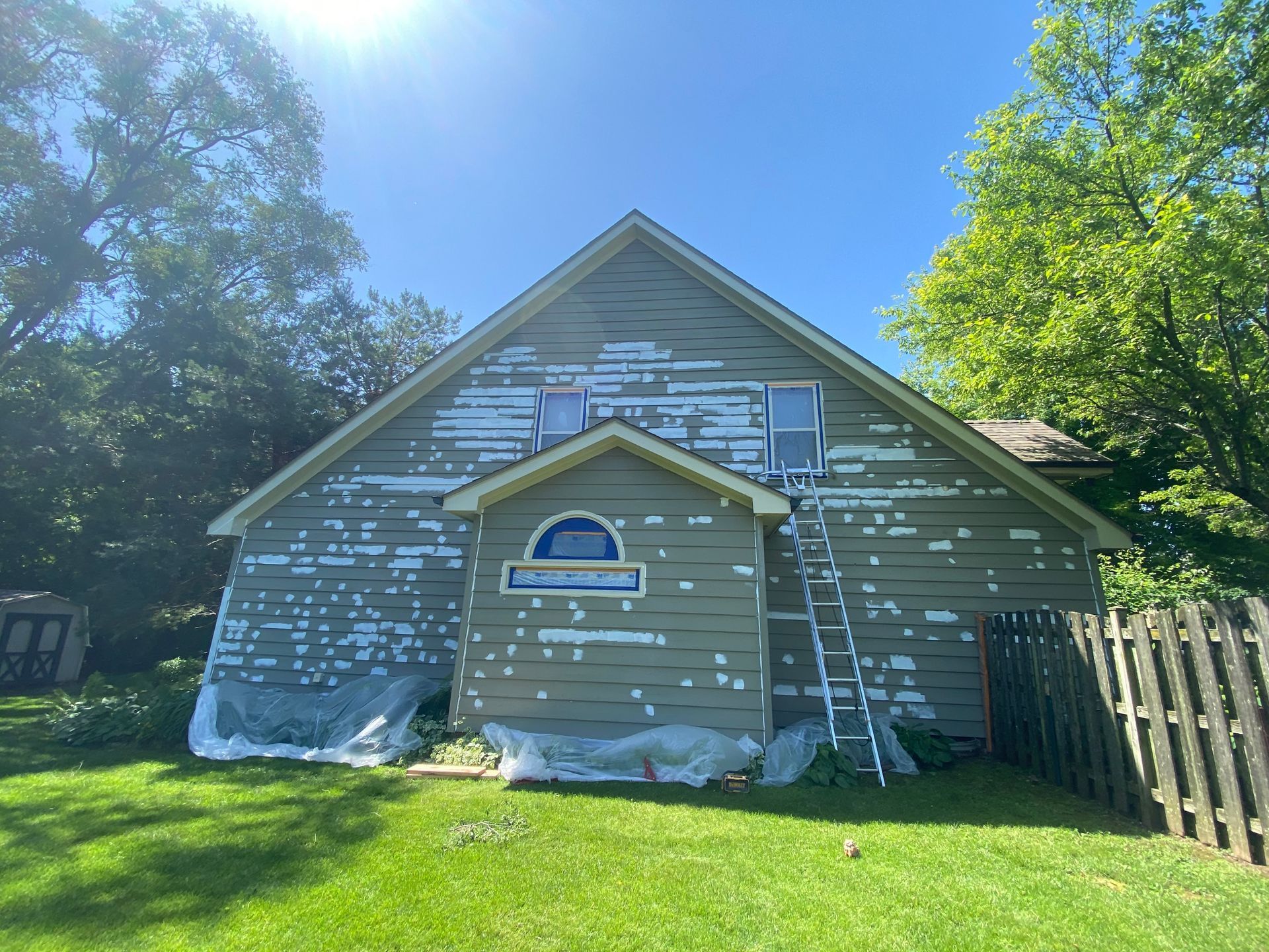 A house exterior with tan, peeling siding, a central arched window, and an extension ladder leaning against the side.