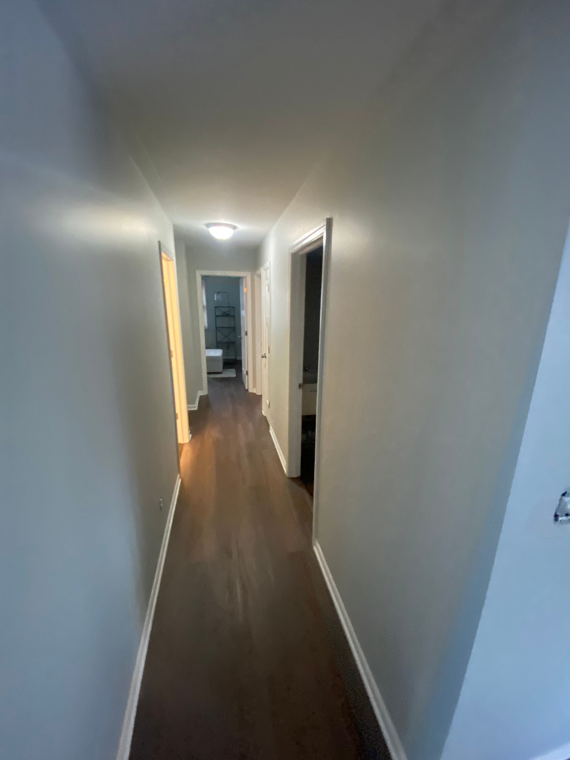 A narrow residential hallway with grey walls and dark wood-look flooring, leading toward two open doorways.
