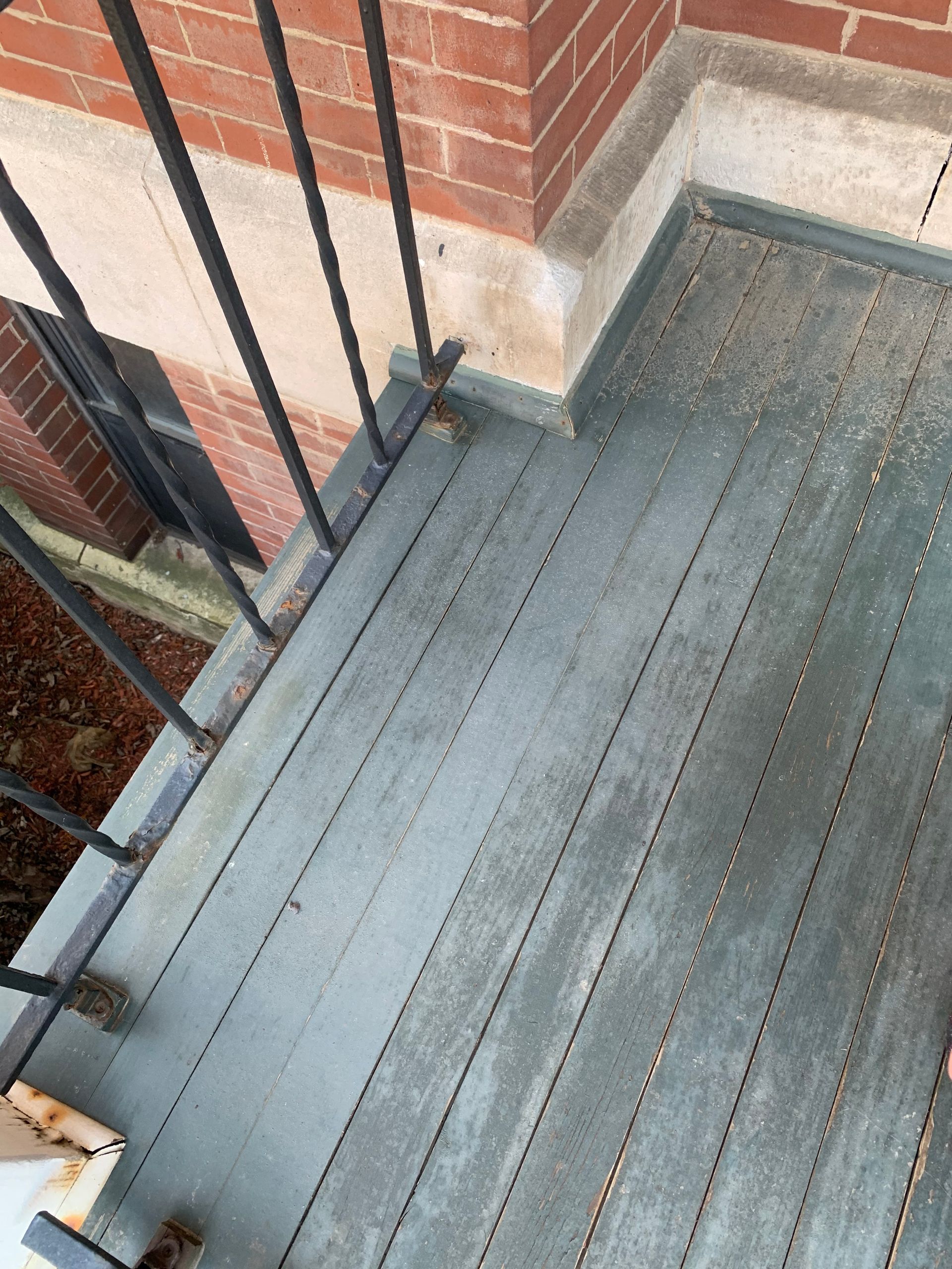 A gray wooden deck with vertical slats, bordered by a black metal railing and a brick wall.