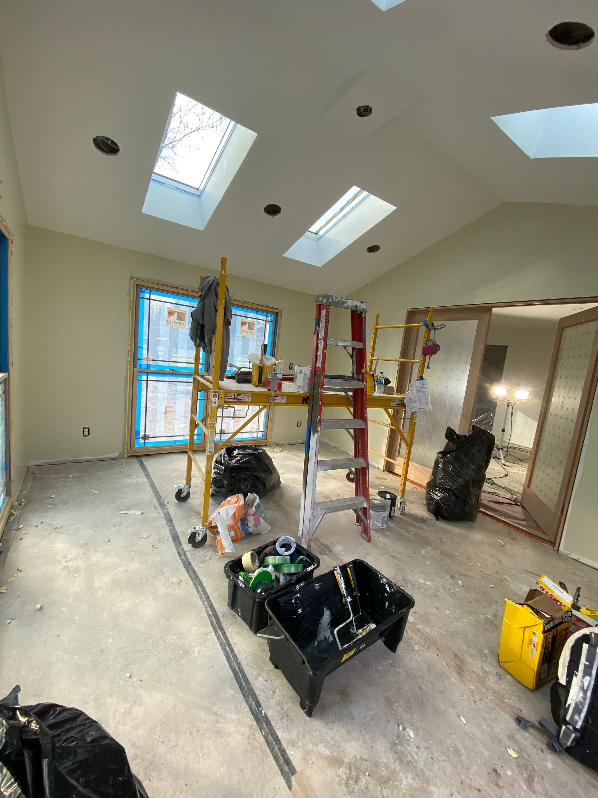 A room under renovation with a metal scaffold, a red step ladder, skylights, paint supplies, and construction debris.