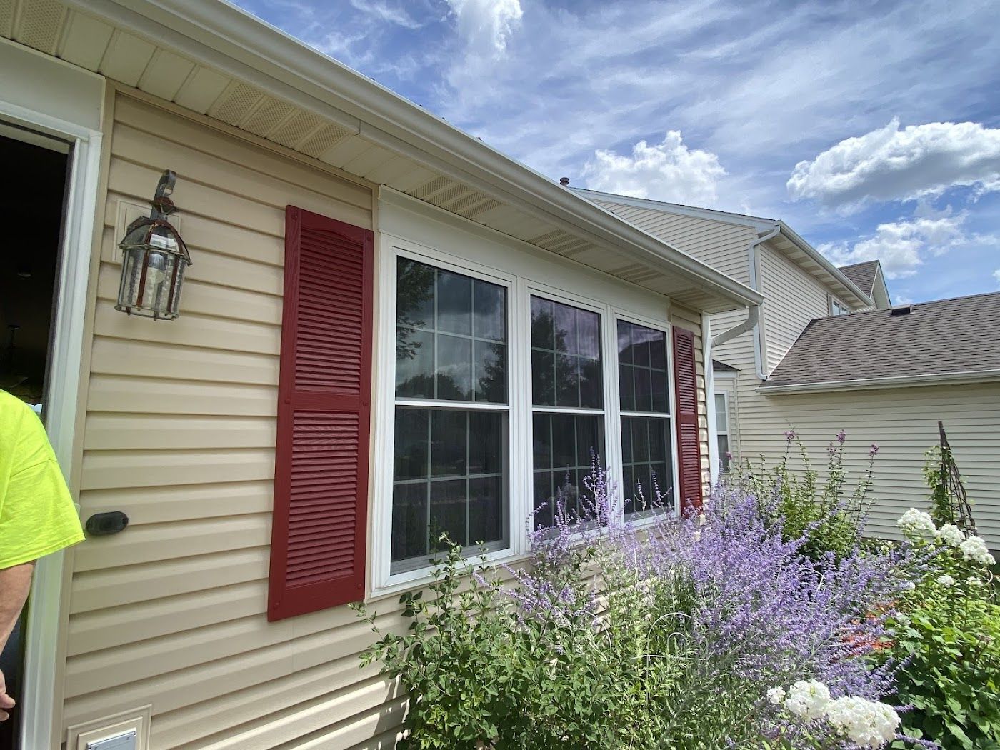 A side view of a beige house exterior with red shutters on a large window and a blooming purple plant in the foreground.