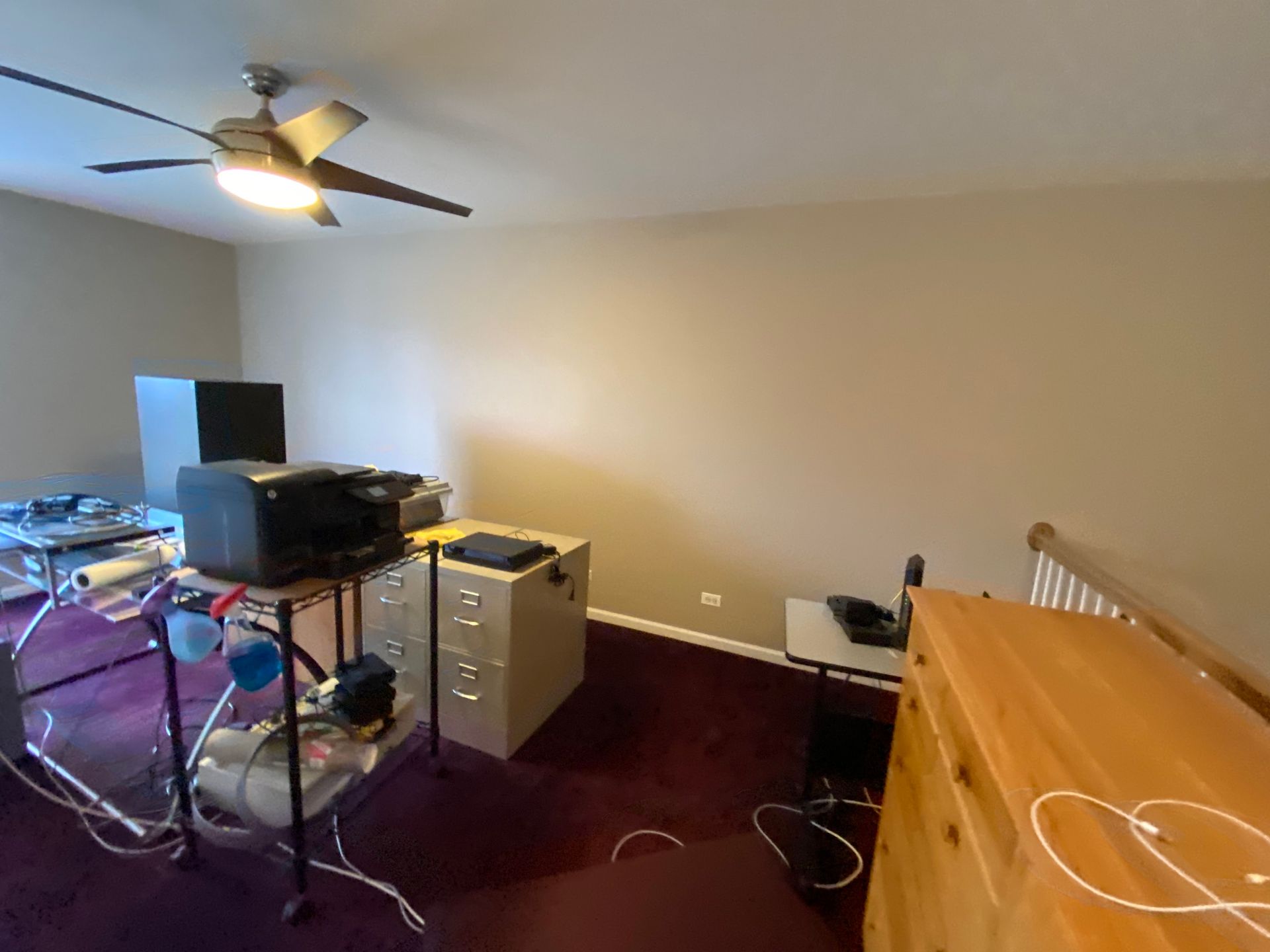 A home office space with a computer setup on a cart, a metal filing cabinet, a wooden dresser, and purple carpet.
