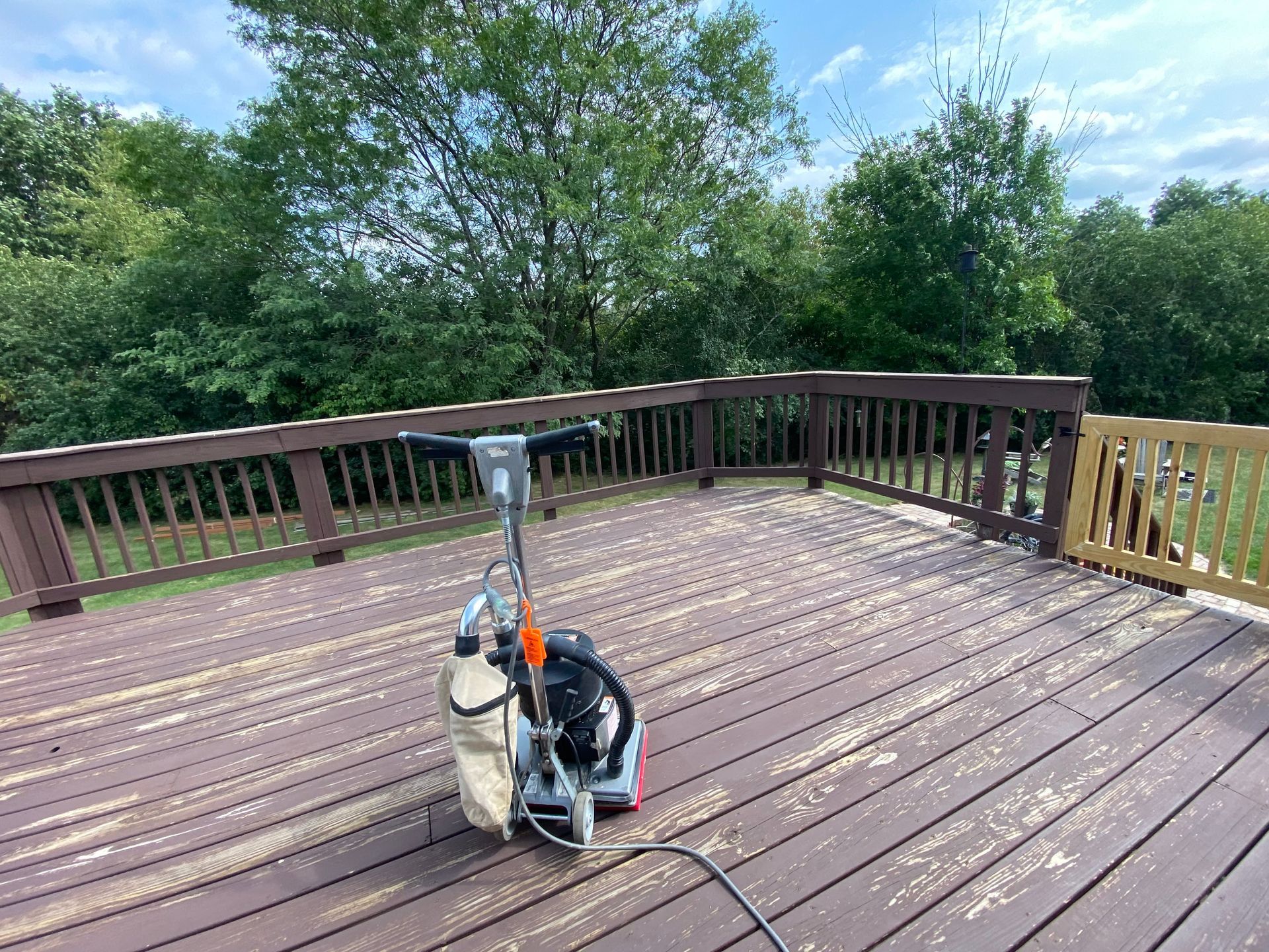 A floor sander stands in the center of a brown wood deck partially stripped of its dark paint, set against leafy trees.