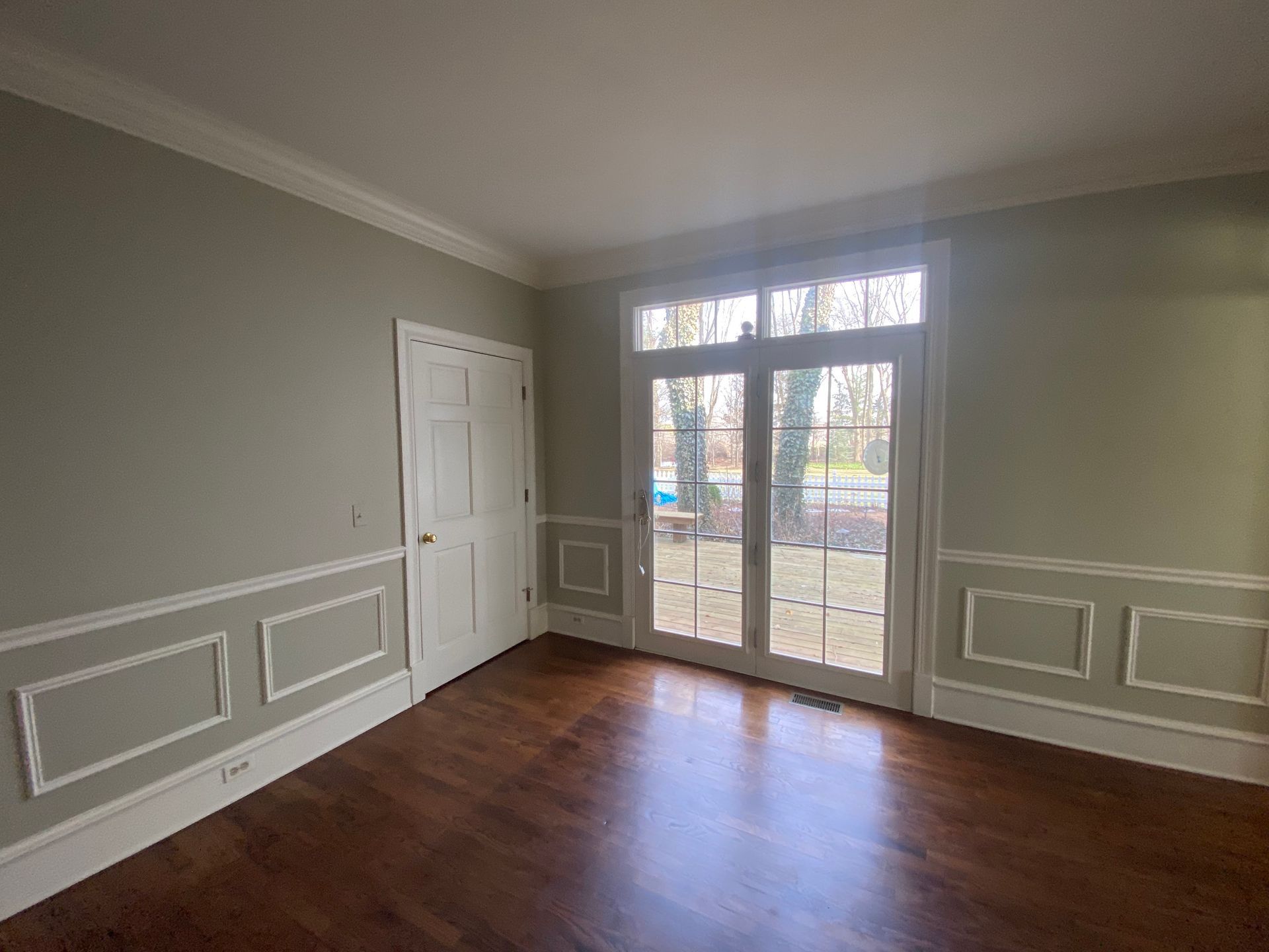 A room with light green walls, white wainscoting, a white door, and glass French doors looking out to a patio and yard.