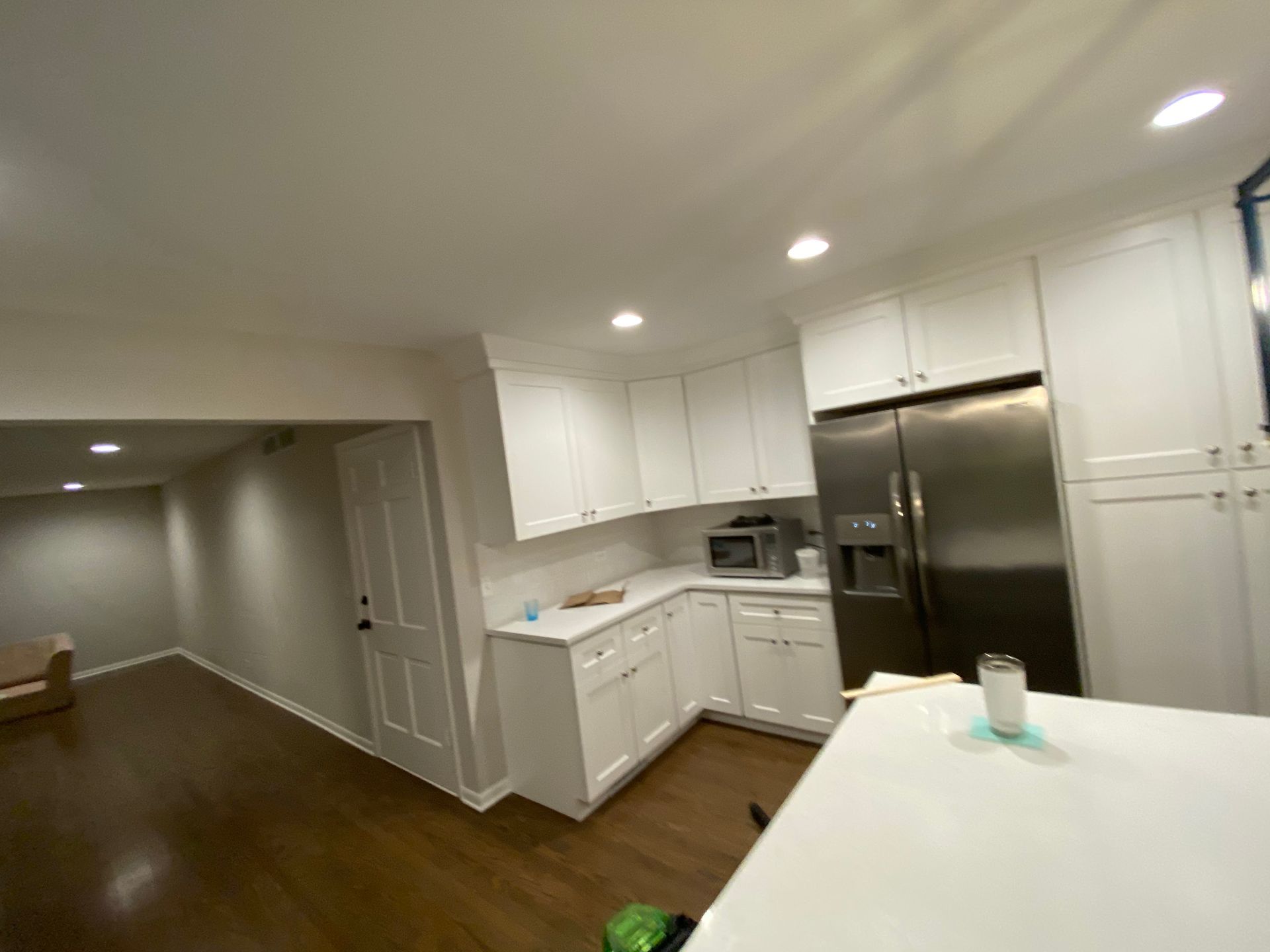 A modern kitchen with white cabinets, stainless steel appliances, and dark hardwood flooring, leading to an empty room.