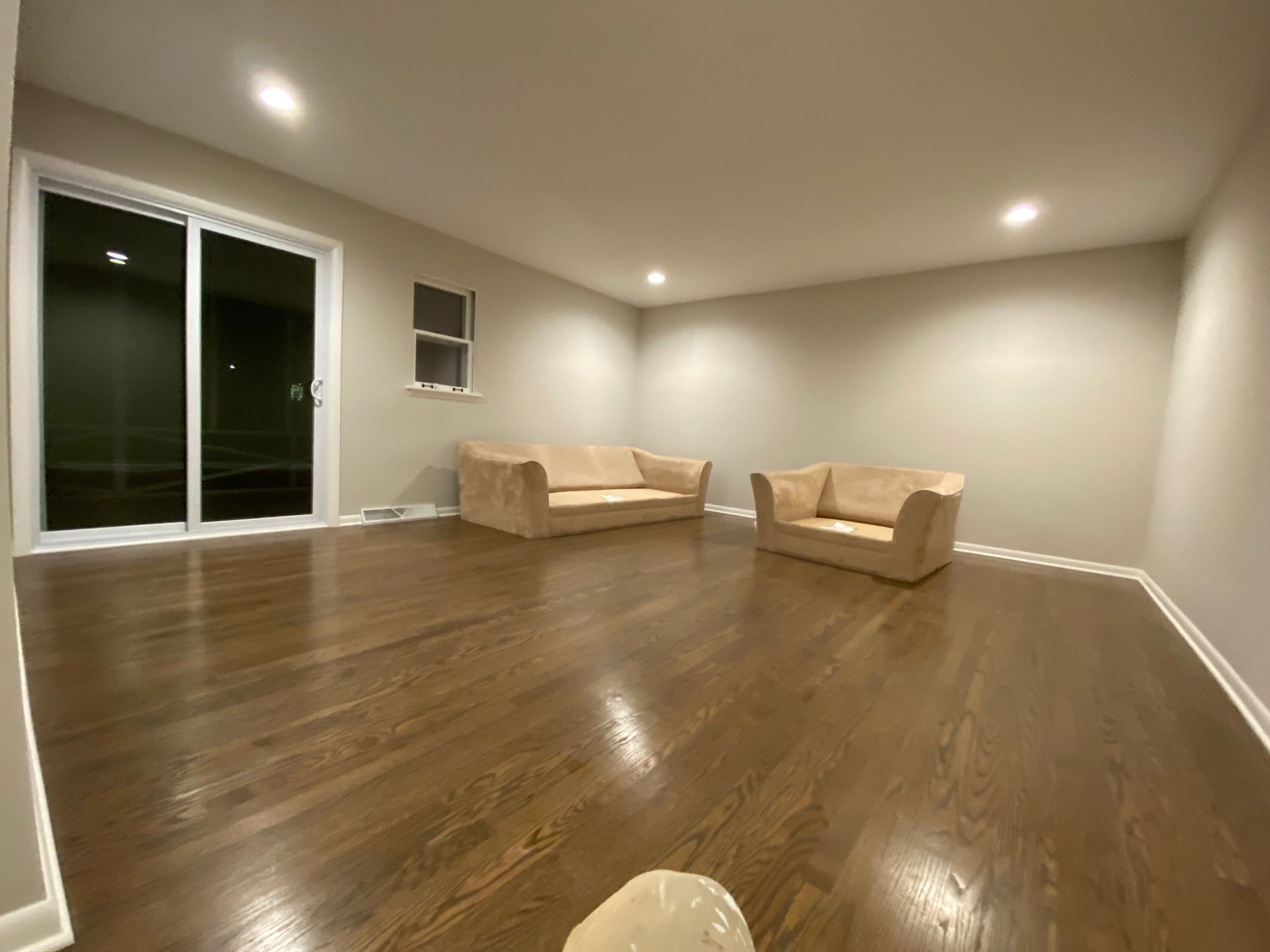A spacious, empty room with dark hardwood floors, beige walls, a sliding glass door, and two tan couches in the corner.
