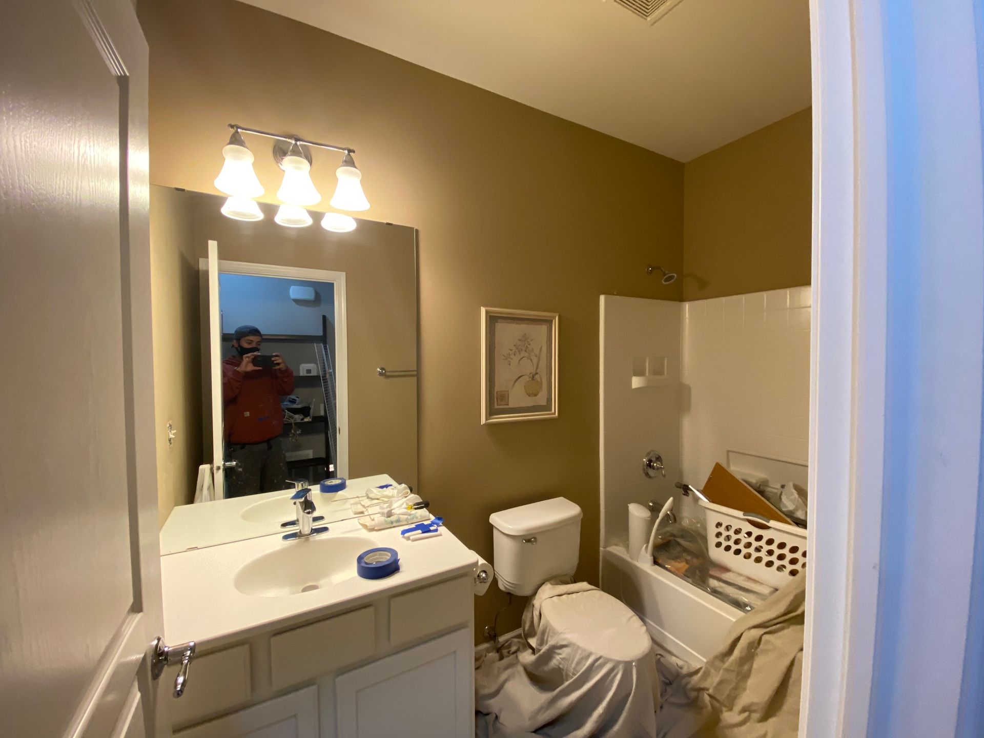 A bathroom with tan walls, a white vanity sink, a toilet with a cloth cover, and a bathtub containing laundry items.