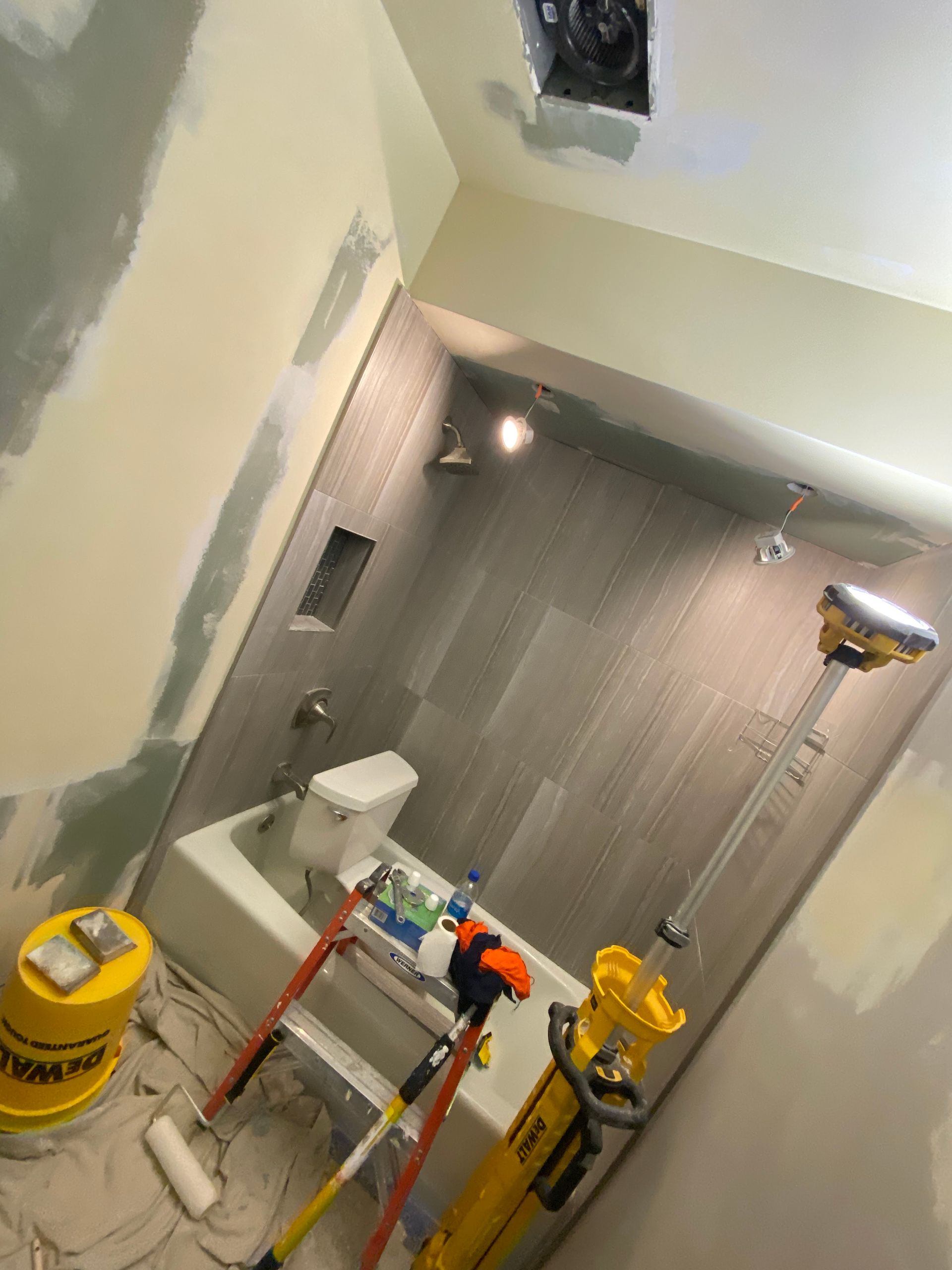 A bathroom under renovation with light gray tiled shower walls, a bathtub, and drywall patching tools on the floor.