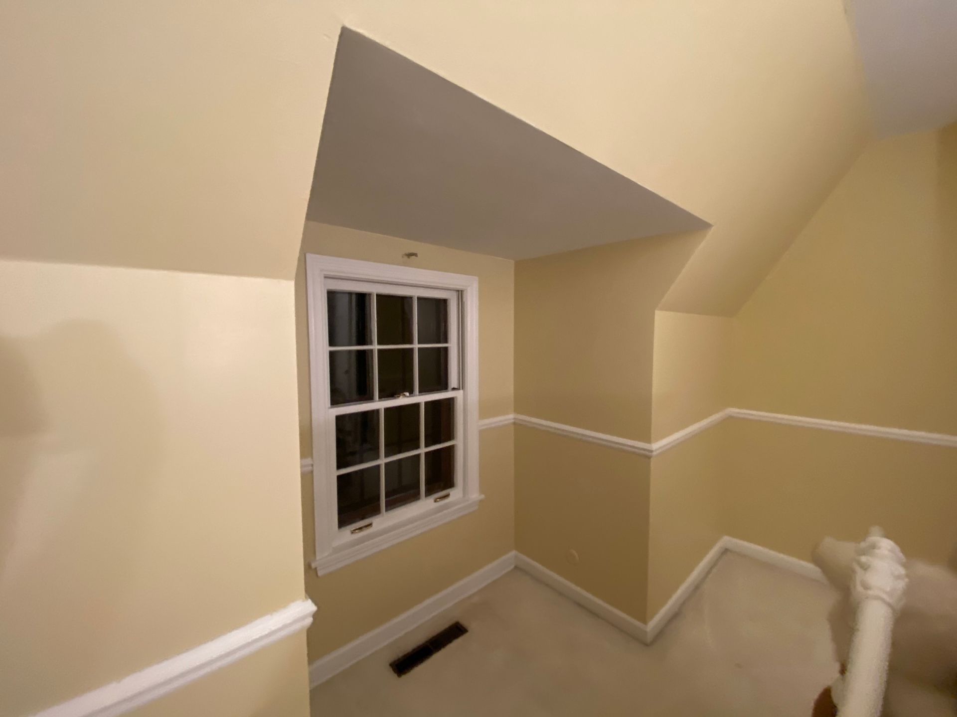 A corner of a room with yellow walls, white trim, a single window, a floor vent, and a sloped ceiling.