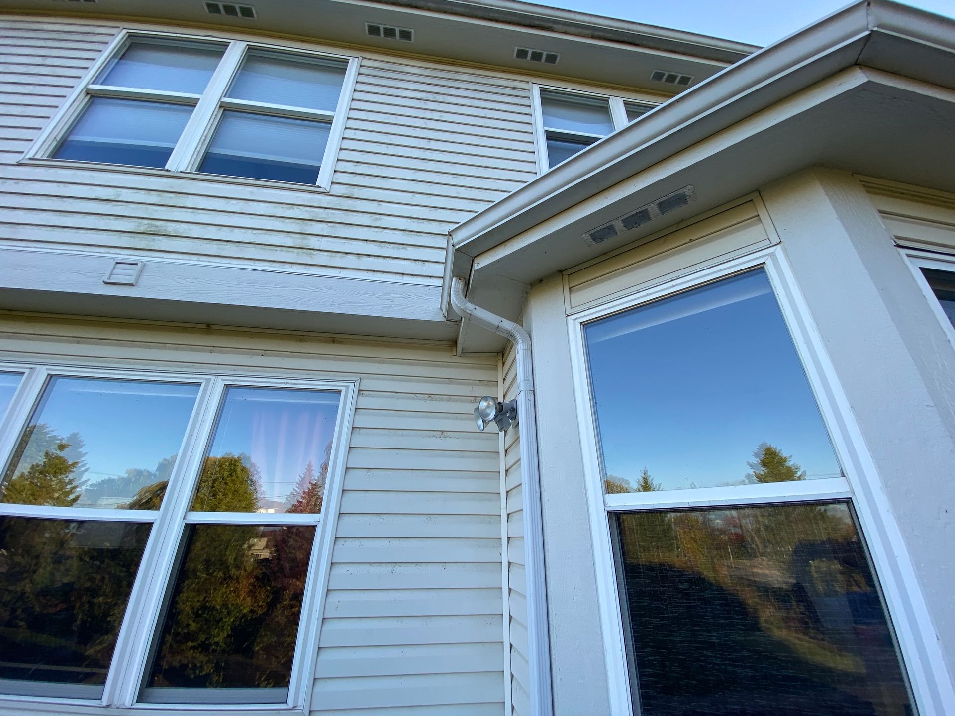 Low-angle view of a light-colored, two-story house exterior with vinyl siding and large windows against a blue sky.