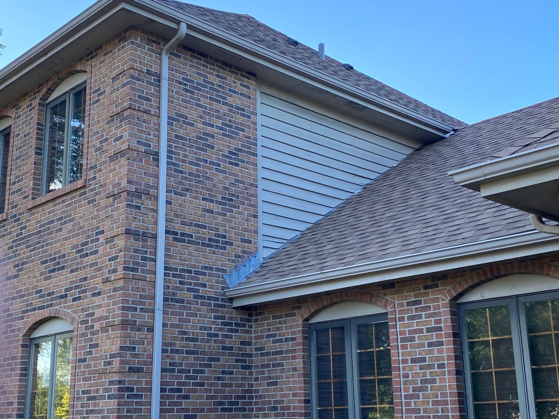 A two-story house exterior featuring tan and dark brick walls, light gray horizontal siding, and dark-framed windows.