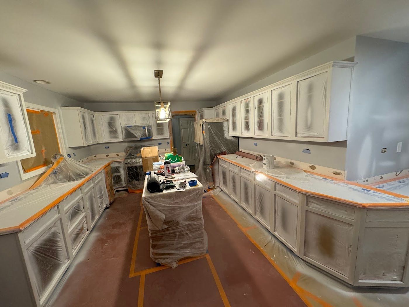 A kitchen undergoing renovation with cabinets masked with tape and plastic sheeting during a painting project.