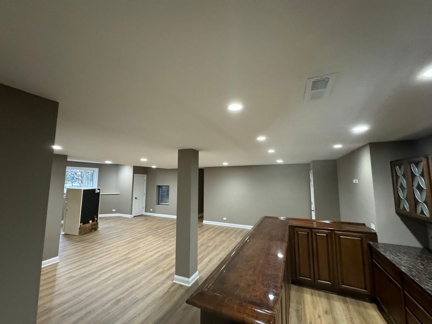 A view of a basement interior with light wood-style flooring, taupe walls, recessed lighting, and a dark wood bar.