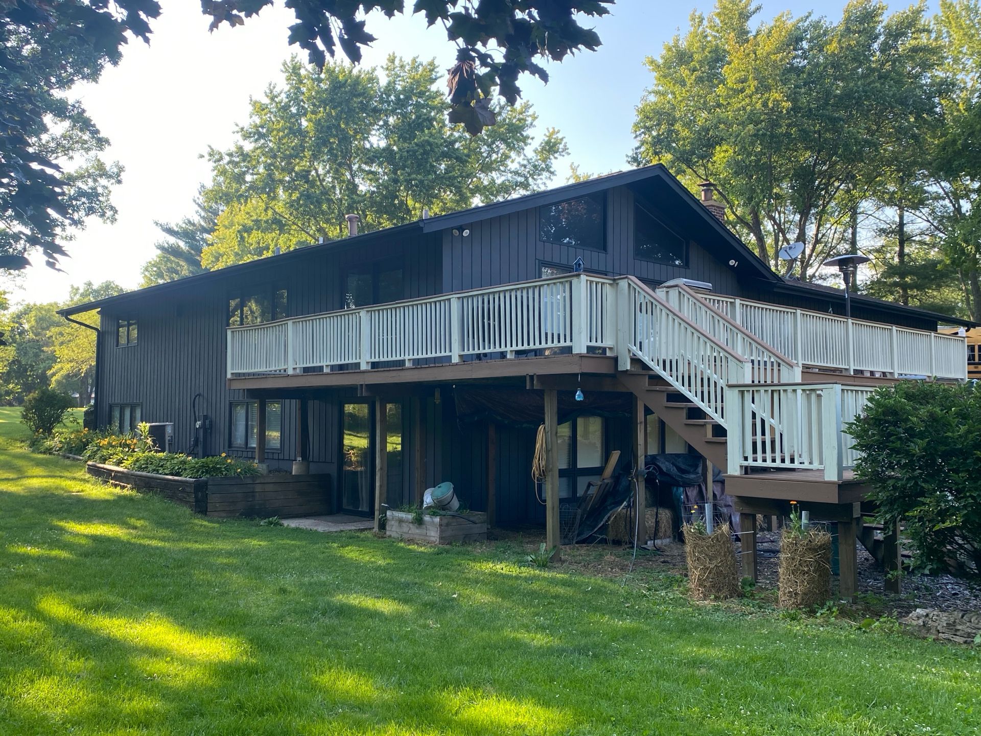 A dark-sided, two-story house with a large white-railed deck extending from the second level over a grassy lawn.